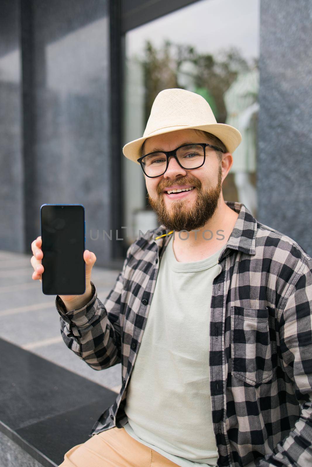 Happy bearded man showing blank smartphone screen over urban background - empty space for advertising and copy space by Satura86