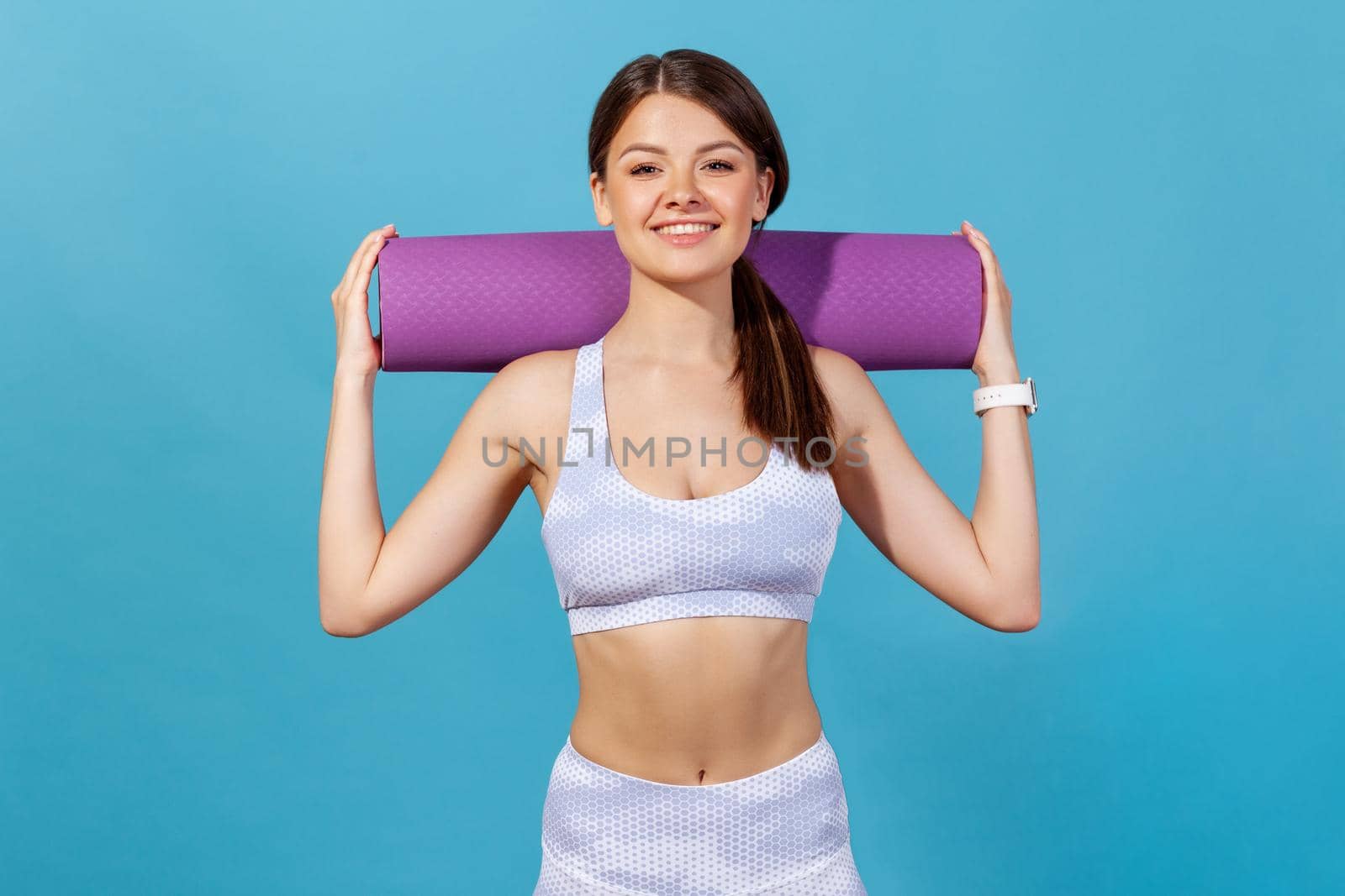 Healthy athletic woman in white sportswear holding rolled mat on shoulders looking at camera with smile, preparing for fitness, pilates or yoga classes. Indoor studio shot isolated on blue background