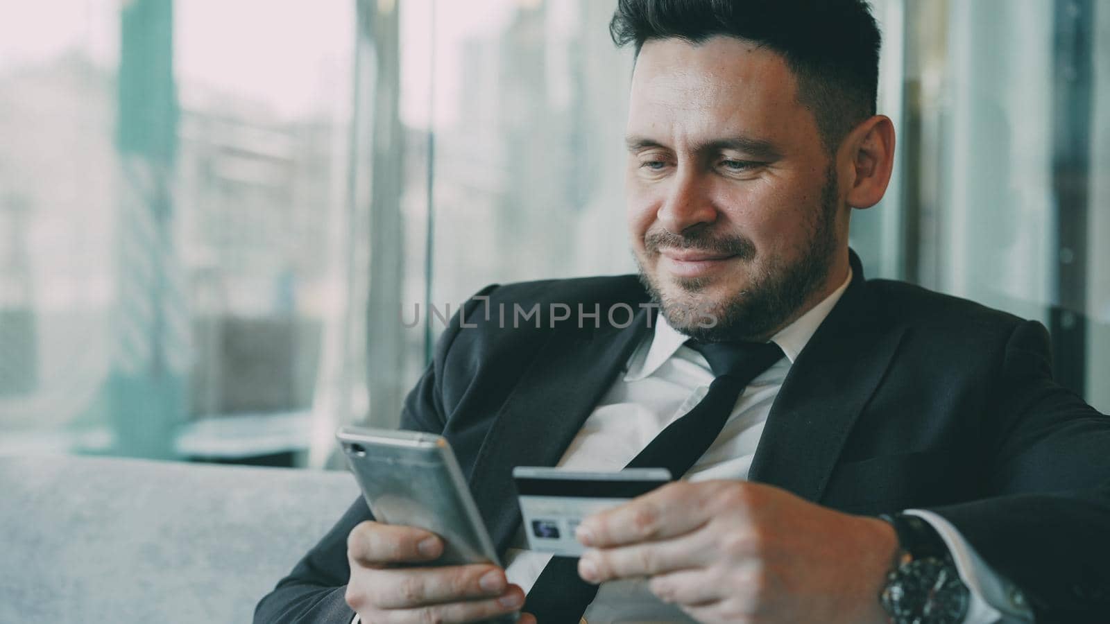 Portrait of cheery bearded Caucasian businessman in formal clothes smiling and paying online bill while keeping credit card and smartphone in his hands in glassy cafe during lunch break.