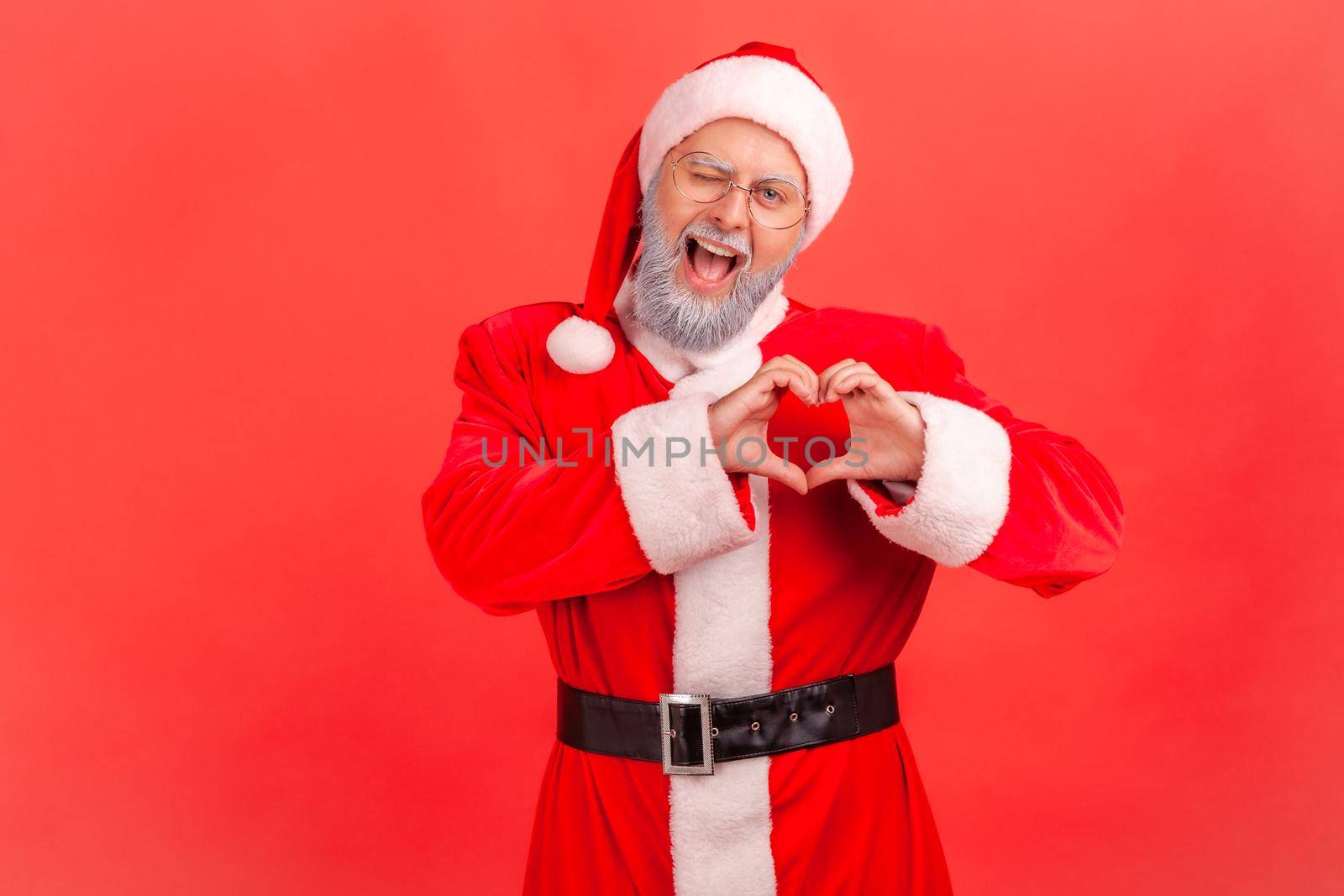 Portrait of friendly positive santa claus in eyeglasses with gray beard showing heart gesture with hands and winking at camera, sending love at holidays. Indoor studio shot isolated on red background