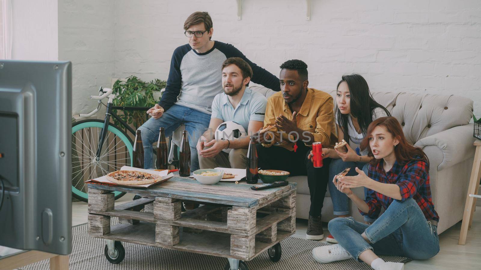 Multi-ethnic group of friends sports fans watching football championship on TV together eating pizza and drinking beer at home by silverkblack