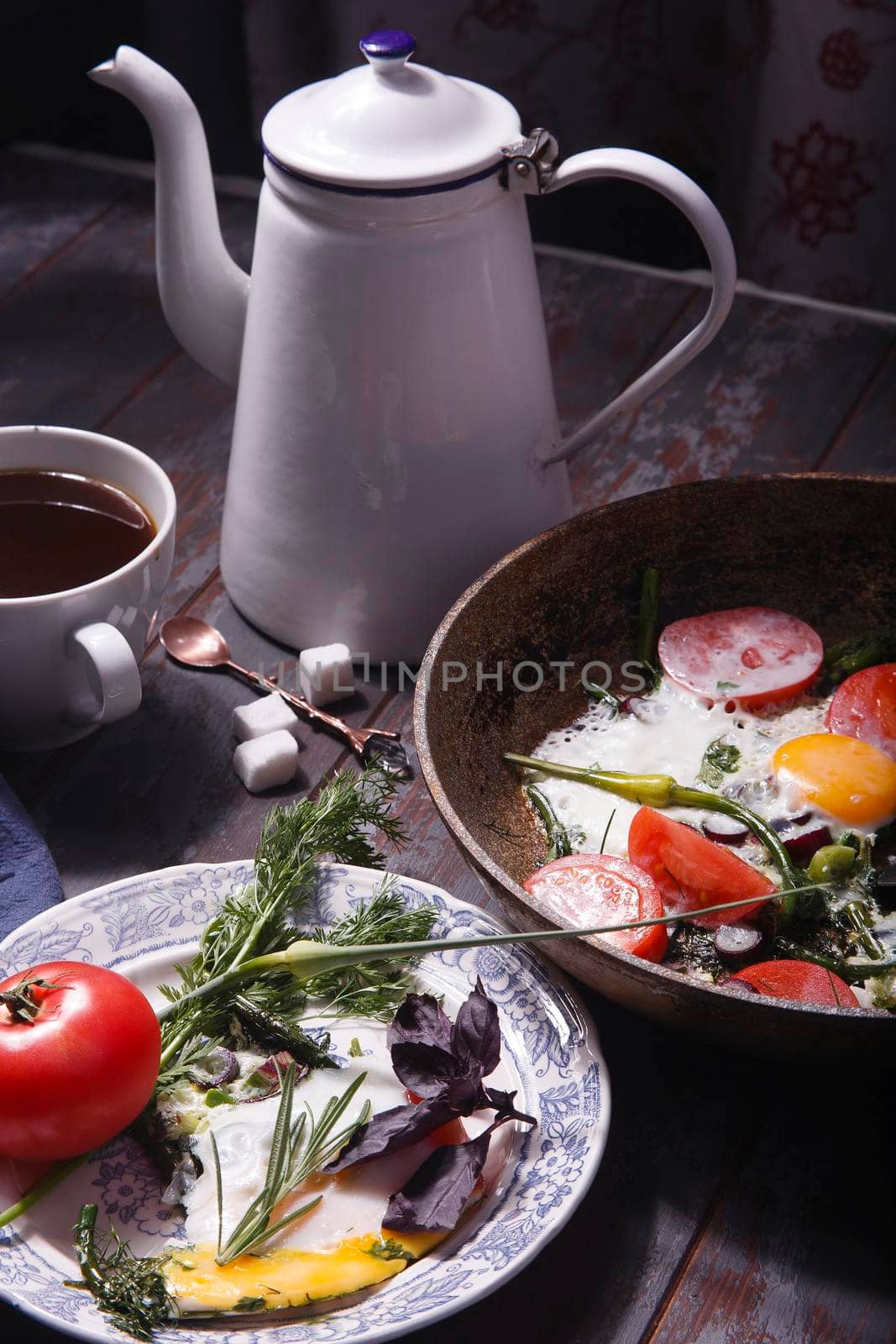 Fried eggs with young garlic stems, greenery, basil, rosemary and tomatoes on frying pan with a cup of coffe and coffe pot, selective focus.