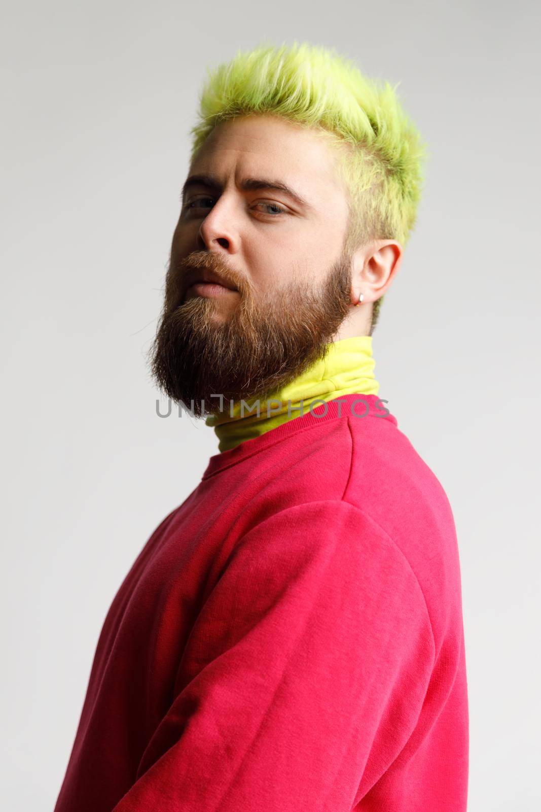 Portrait of proud handsome young bearded Caucasian man in red casual sweater, looking with prideful face,standing half sideways smug puffed. Indoor studio shot isolated on gray background.