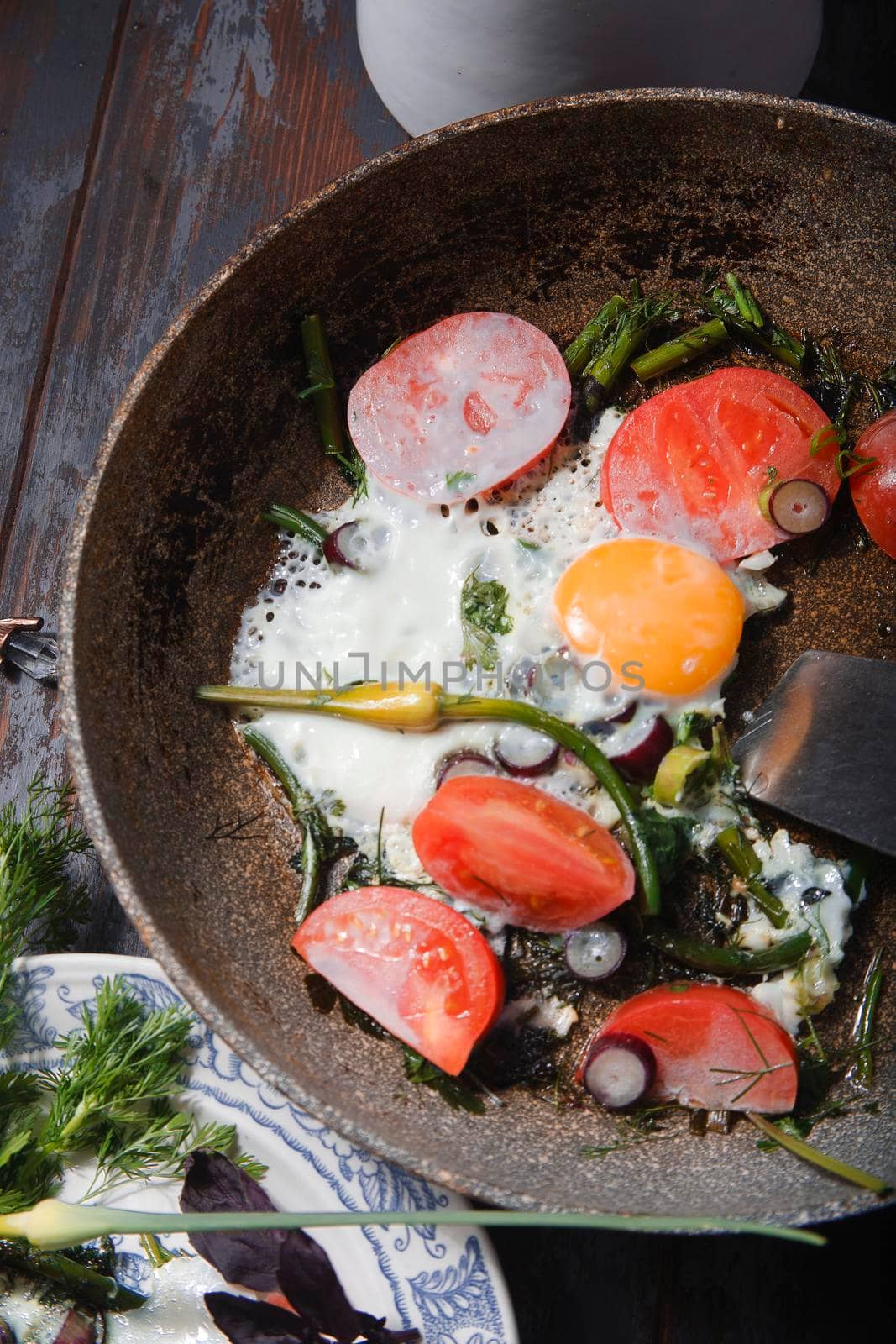 Summer breakfast. Fried eggs with young garlic stems, greenery, basil and tomatoes on frying pan, close up, flat lay.