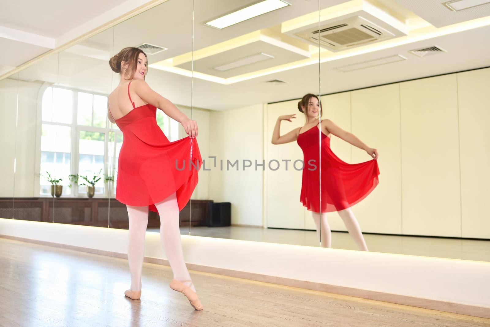 a young ballerina in a red dress and pointe shoes performs exercises in a dance studio