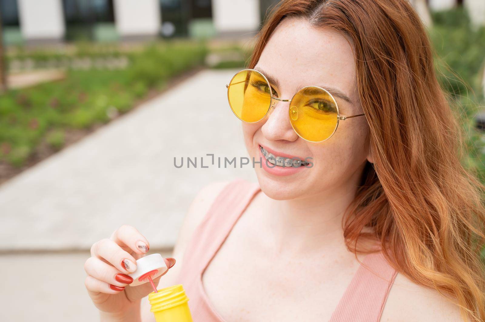 Young red-haired woman blowing soap bubbles outdoors. Girl in yellow sunglasses and braces