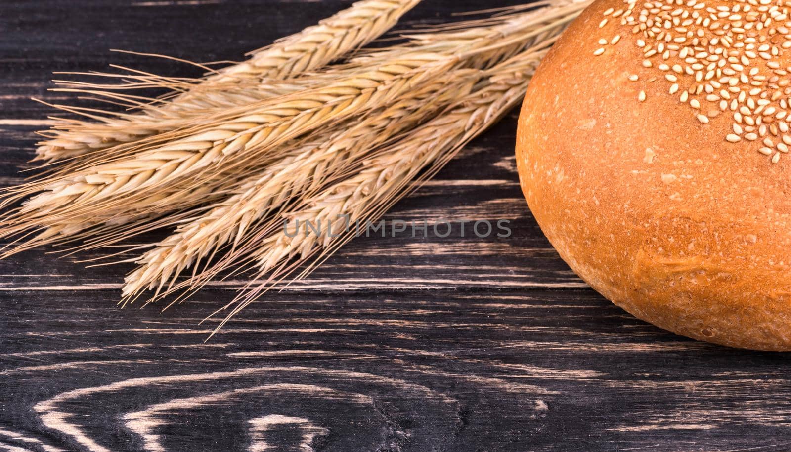 The fluffy bun with sesame seeds and spikelets of wheat on dark wooden background