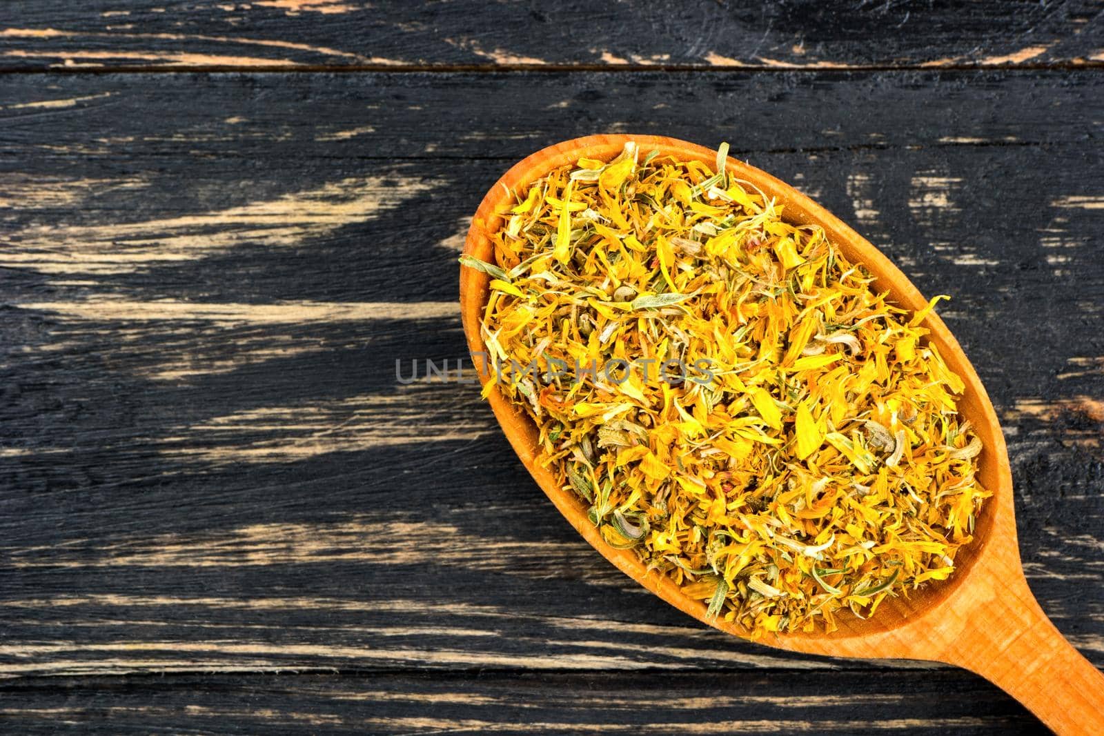 Wooden spoon full of dried calendula flowers close up on a dark table,  top view