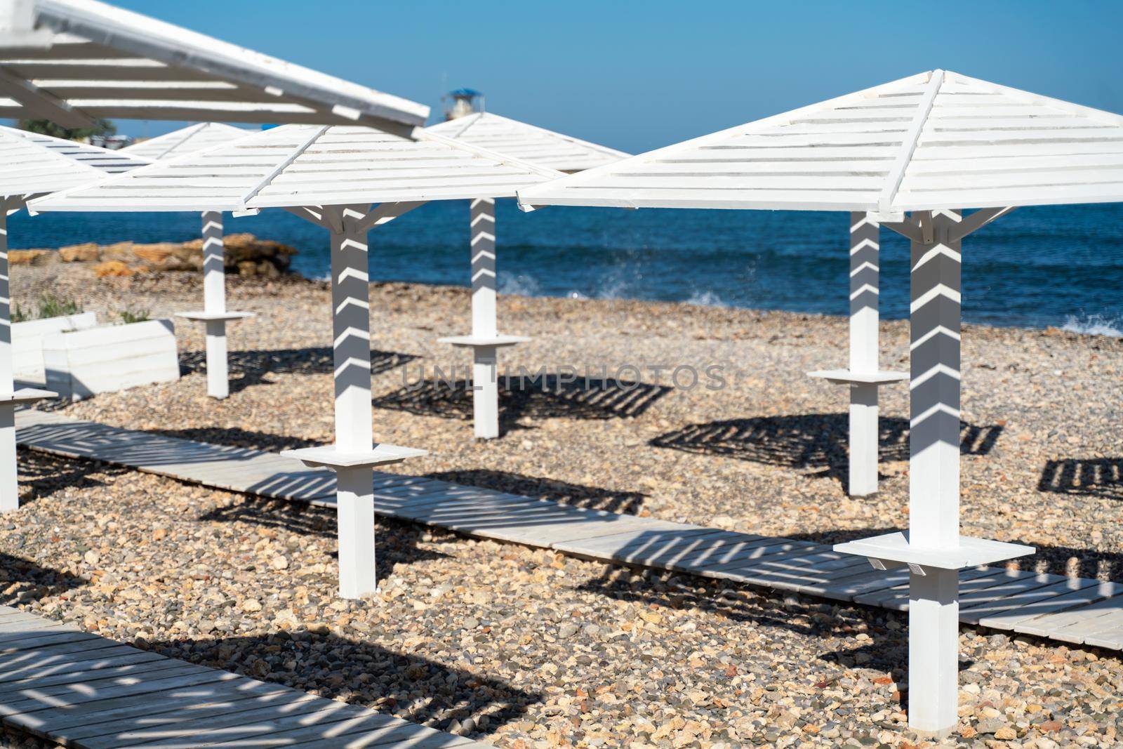 Rows of wooden umbrellas from the sun on the seashore in the morning. Wooden paths on the sand between umbrellas. Beach holiday at the resort. by Matiunina