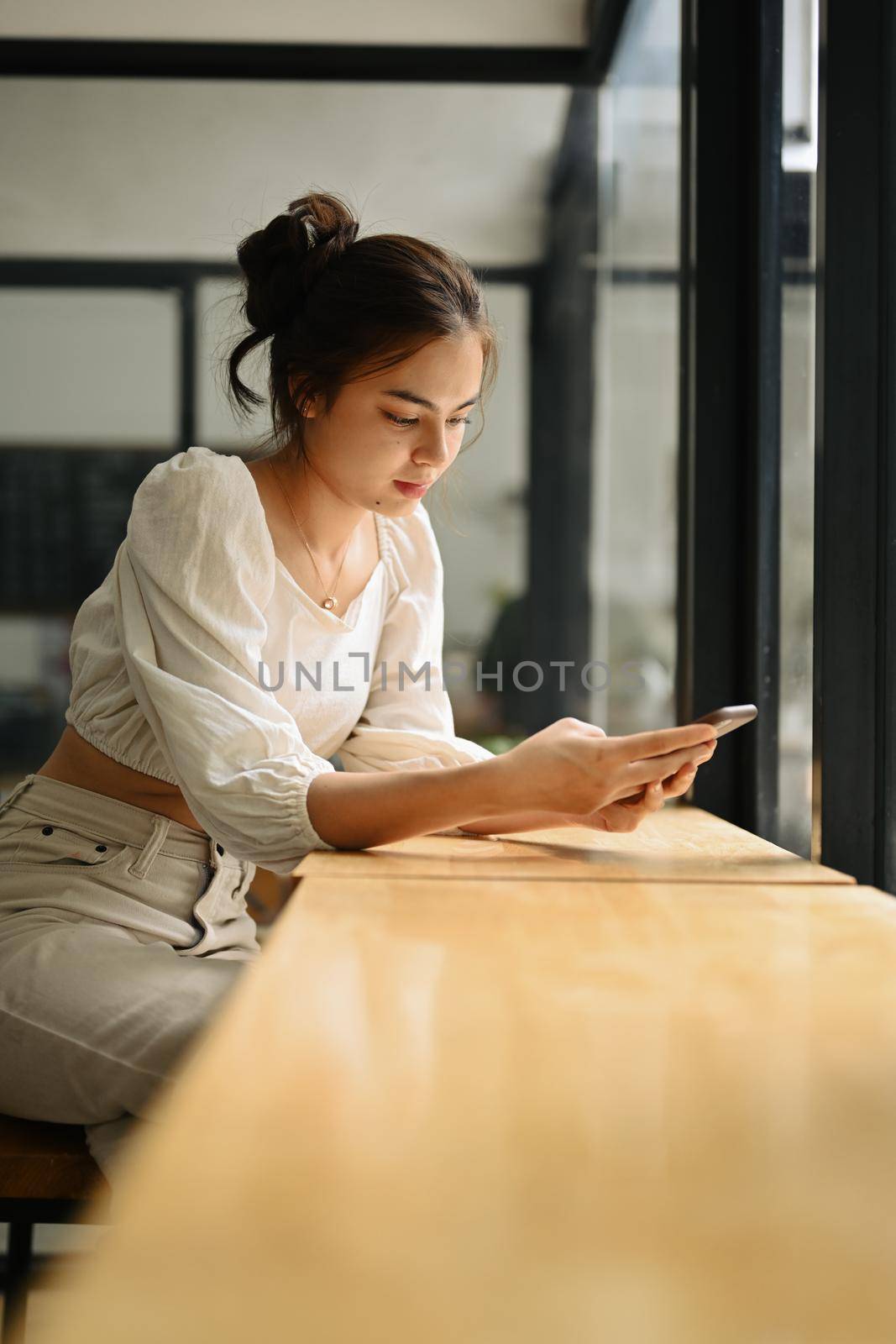 Charming young woman sitting near window in coffee shop and messaging on her smartphone.