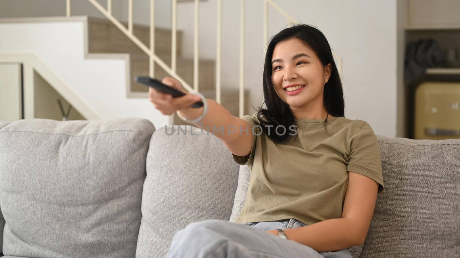 Happy young woman holding remote control, watching tv on couch, enjoying carefree leisure weekend activity at home by prathanchorruangsak