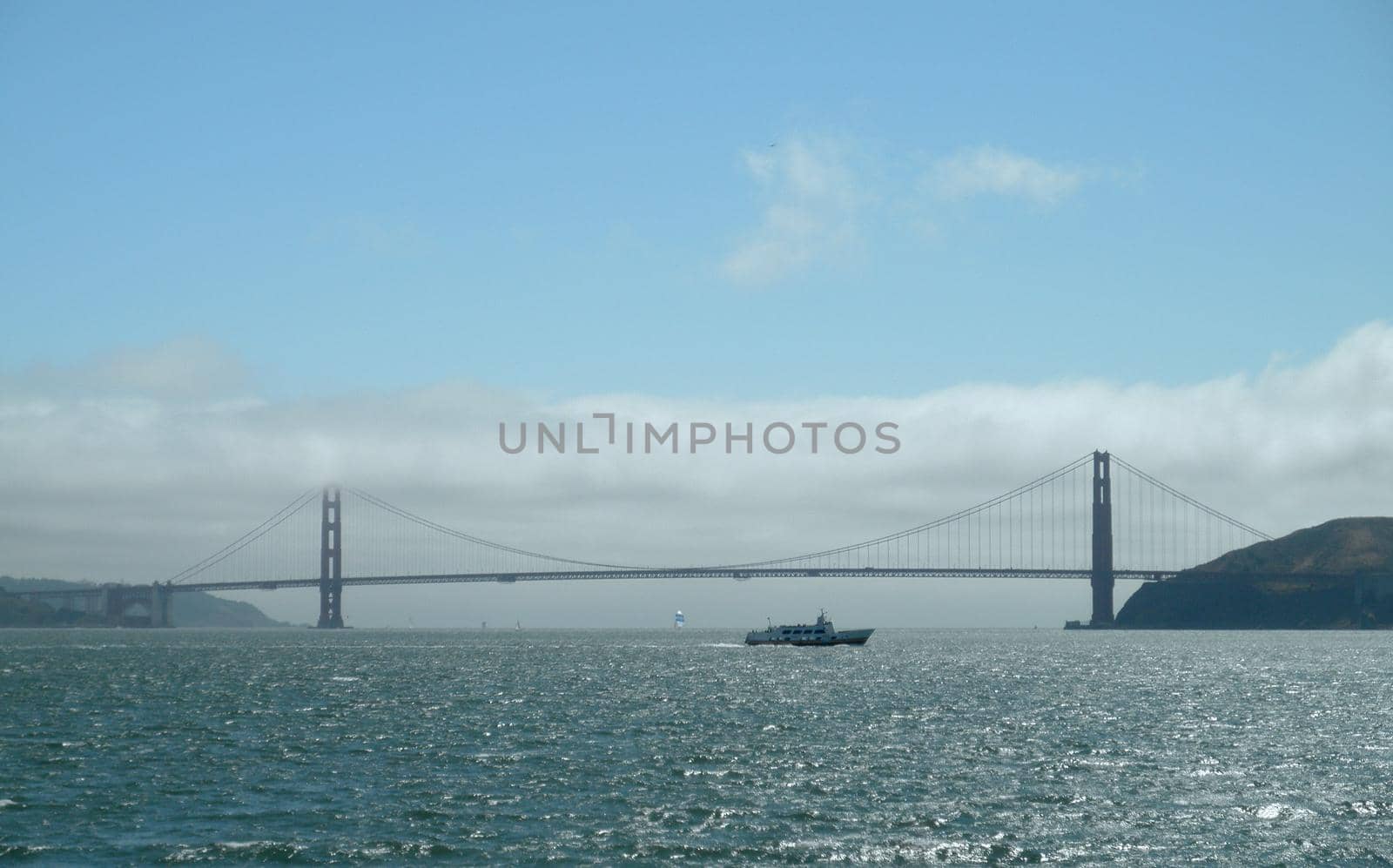 Ferry boat passes the Golden Gate bridge, which is half covered in fog and in full view, on the bayside.  Sailboats can be seen in the distant