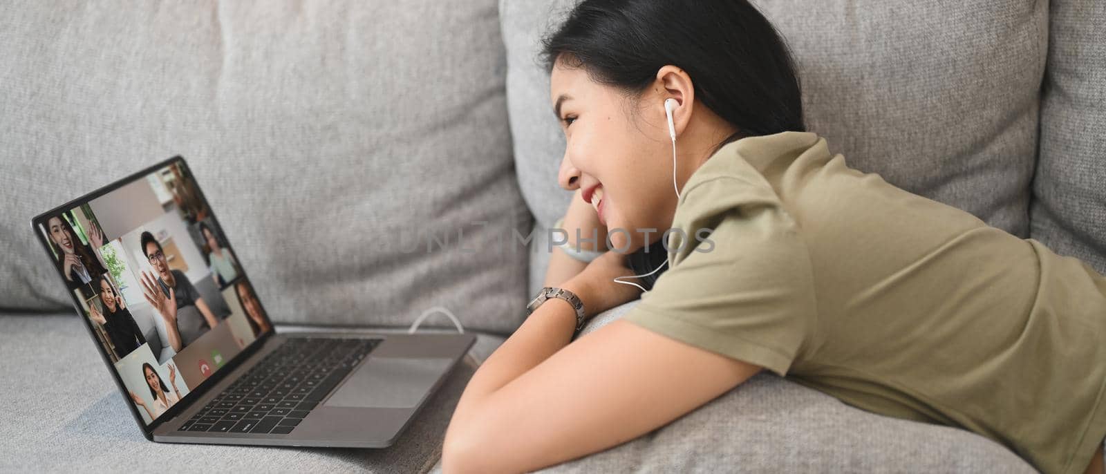 Cheerful young woman communicating by video conference via laptop while lying on couch at home.