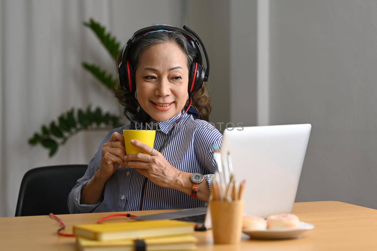 Smiling middle aged woman wearing headphone watching online webinar on laptop computer at home kitchen.