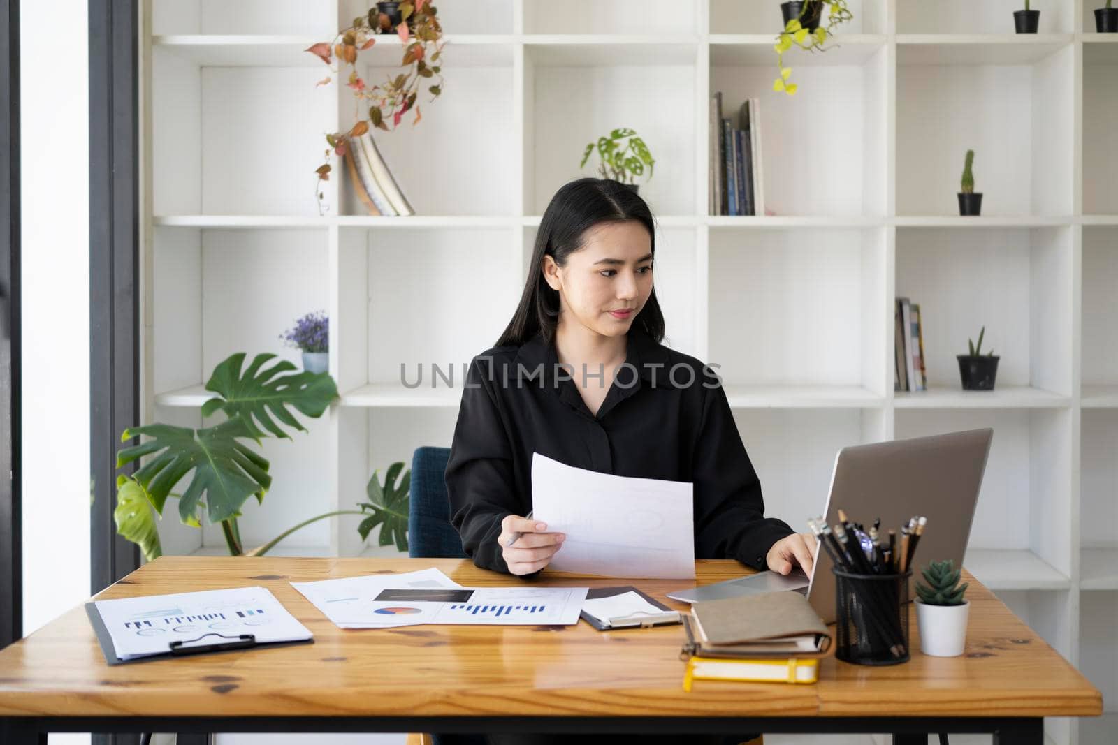 Charming businesswoman holding document and working with laptop at her office desk.