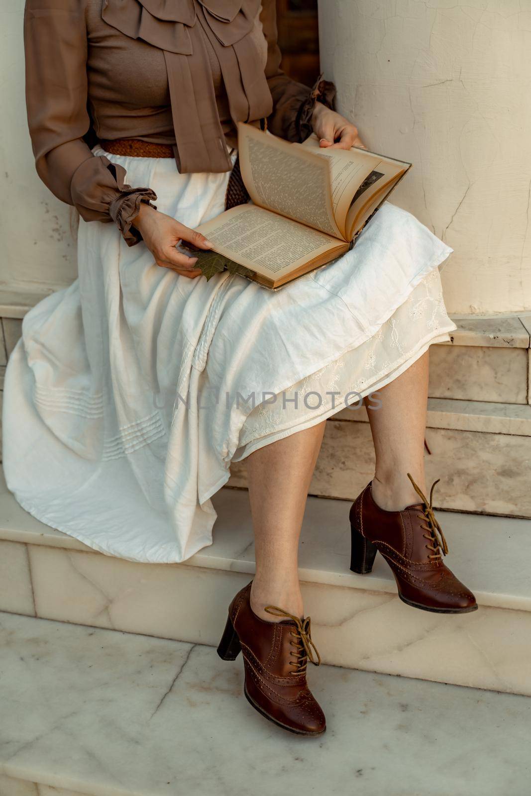 A woman in a white skirt and brown blouse sits and holds an open book in her hands. She is reading a book