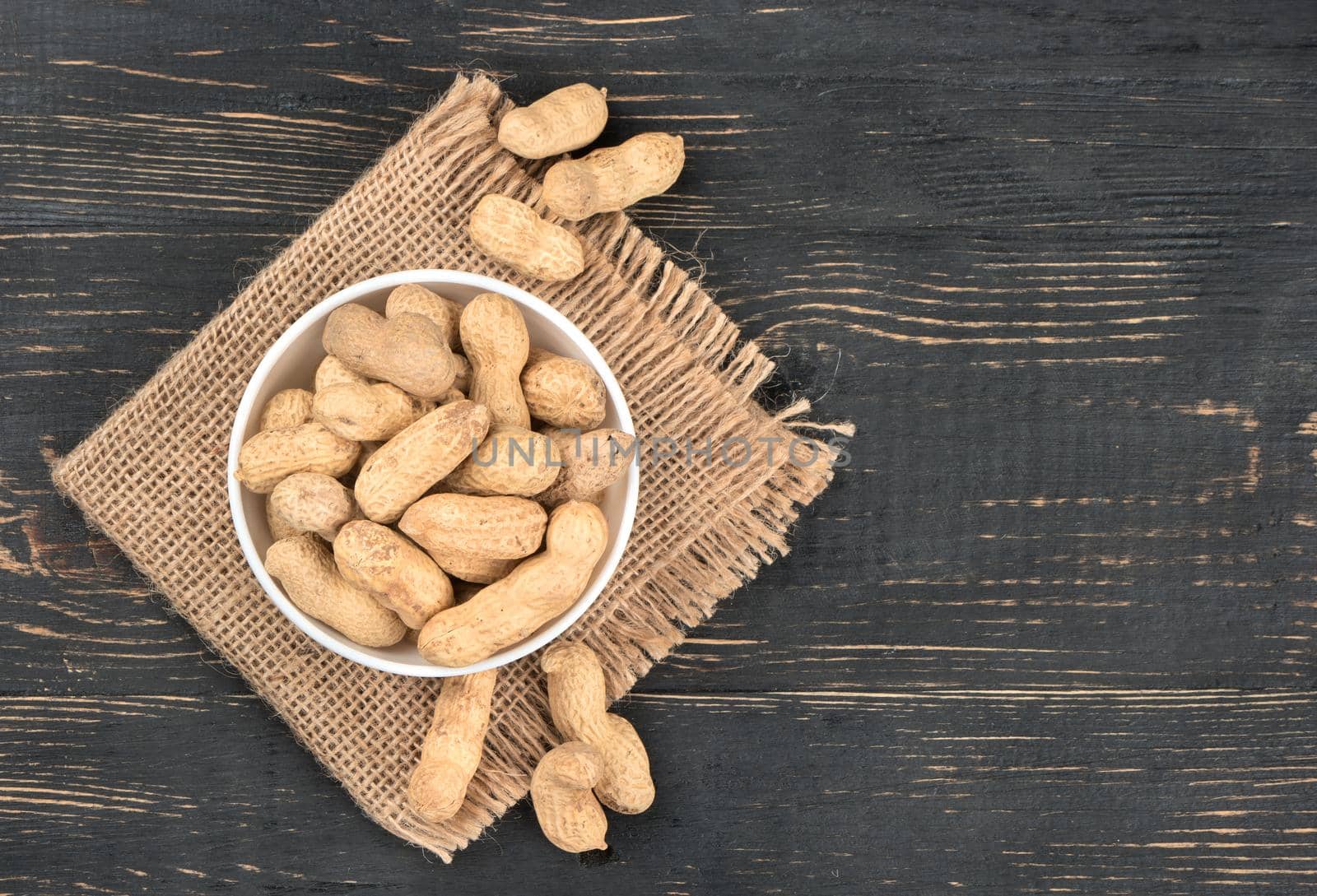 Full ceramic bowl of peanuts in the shell on sackcloth and empty wooden background, top view