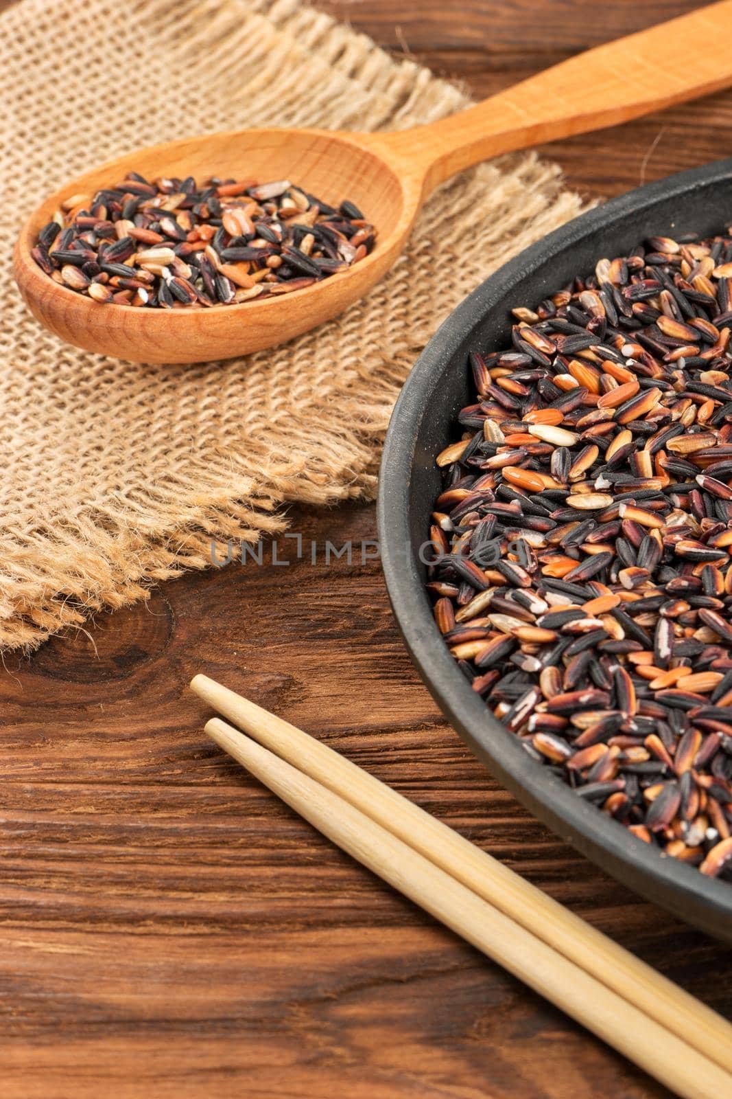 Part of frying pan with black wild rice, chopsticks and spoon on wooden background