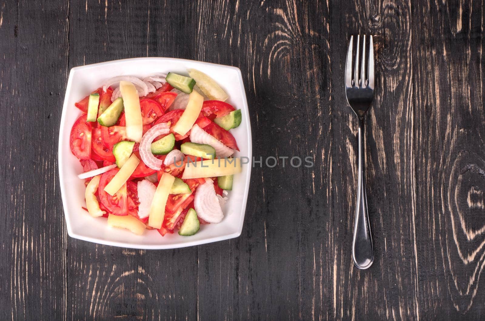 Fresh salad with tomatoes, cucumbers, peppers and onions on a plate with a fork on a dark wooden table