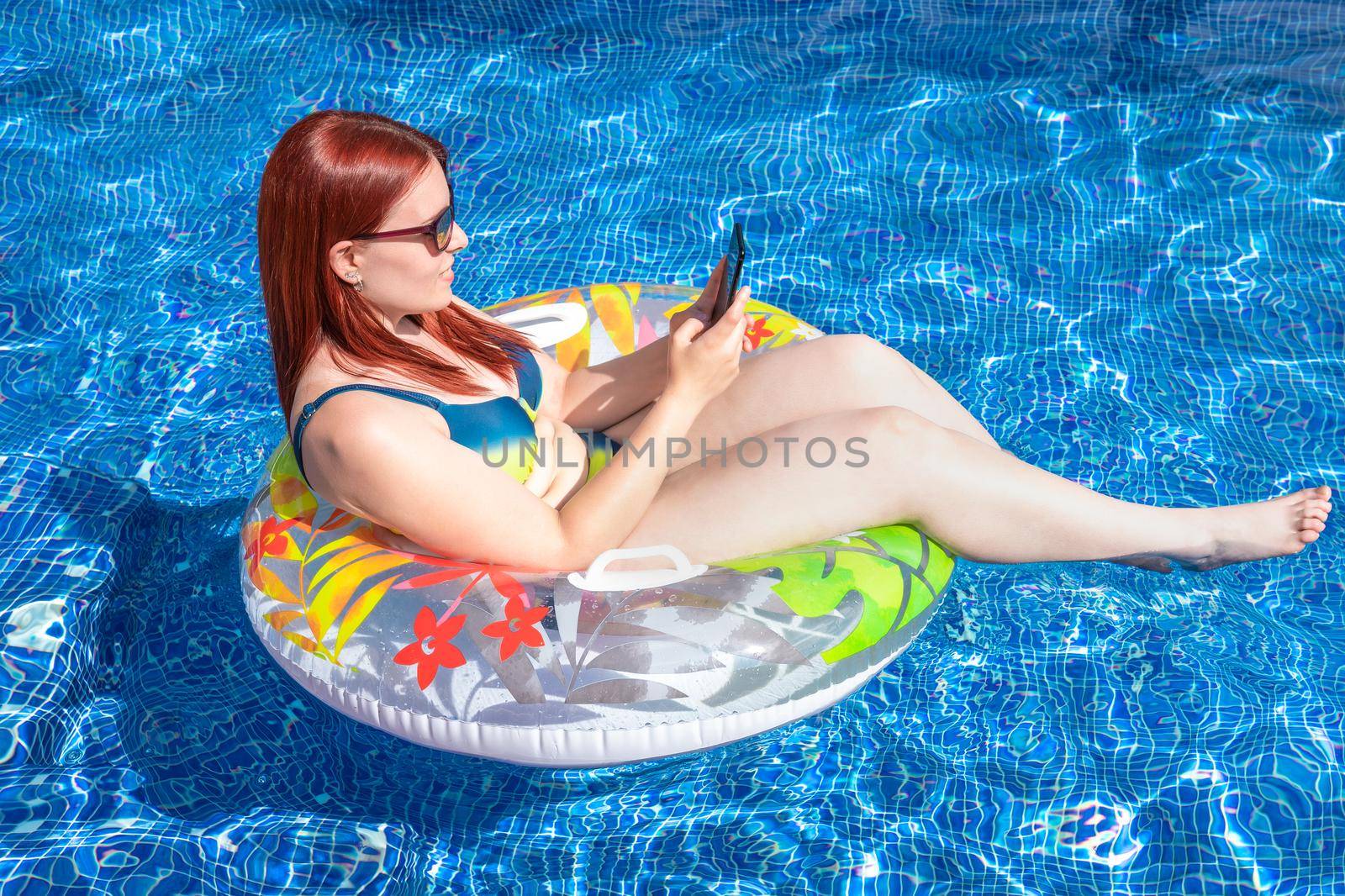 young redhead girl in bikini, looking at her smartphone floating on a ring float inside the pool. young girl on holiday relaxing, sharing the moments on her social networks. by CatPhotography
