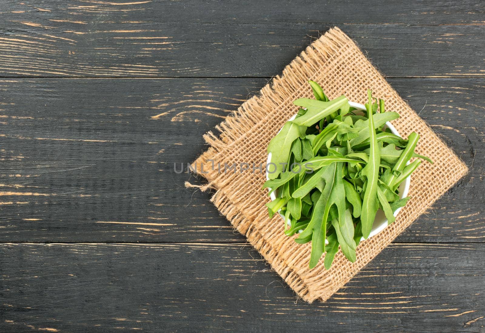 Fresh arugula in bowl on burlap on blank wooden background, top view