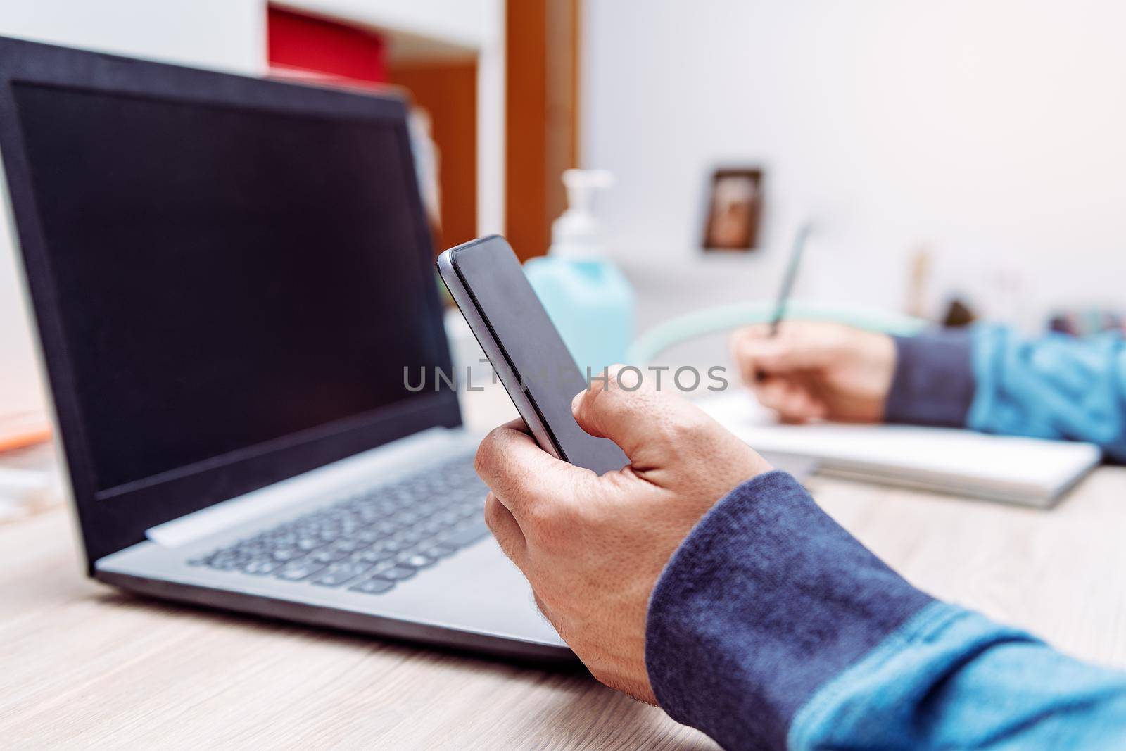 detail of the hand of a man holding a smartphone, out of focus is a computer and a hand sanitizer gel, work at home concept due to the coronavirus pandemic