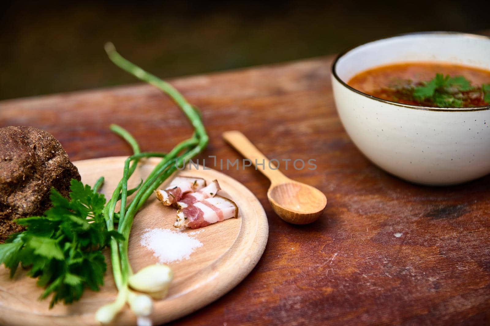 Sliced bacon, green onion, parsley and bread on a wooden board nearby a bowl with Ukrainian traditional dish- Borscht by artgf