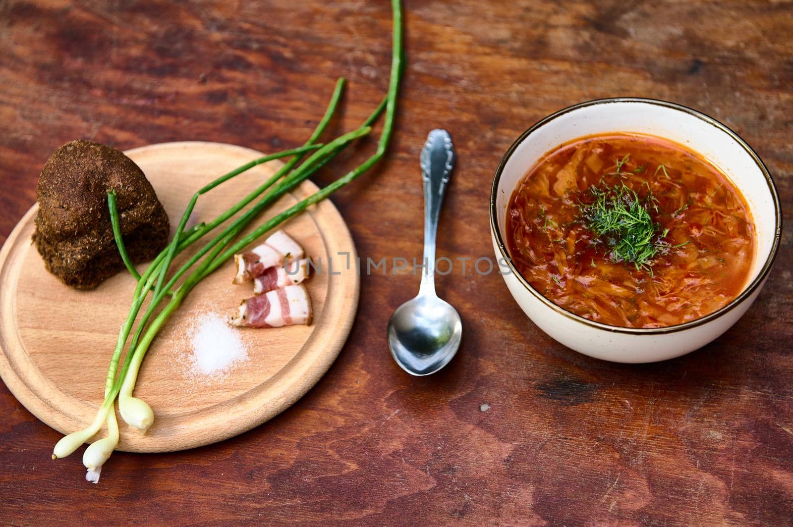 Overhead view of a bowl of beetroot soup with parsley - Ukrainian borscht - and a wooden board with green onion, dill, salt, bacon and whole grain rye bread. Traditional Ukrainian cuisine