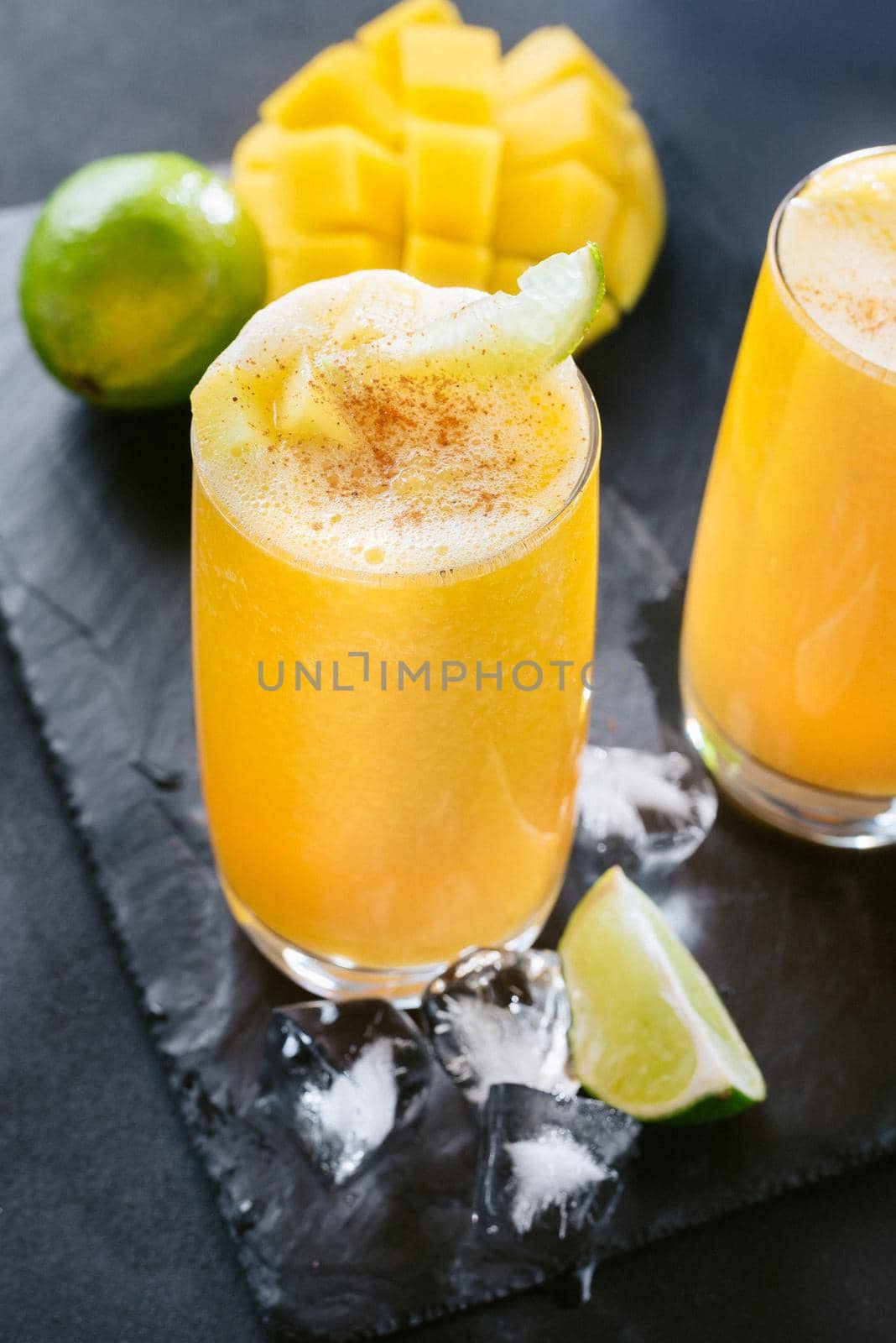 Mango juice on a dark background. Summer drink with ice and slices of mango and lime. Bright yellow summer mango cocktail. Slices of mango and lime on a black background. Seasonal drink. Cocktail for vegetarians and those on a diet. Healthy Food