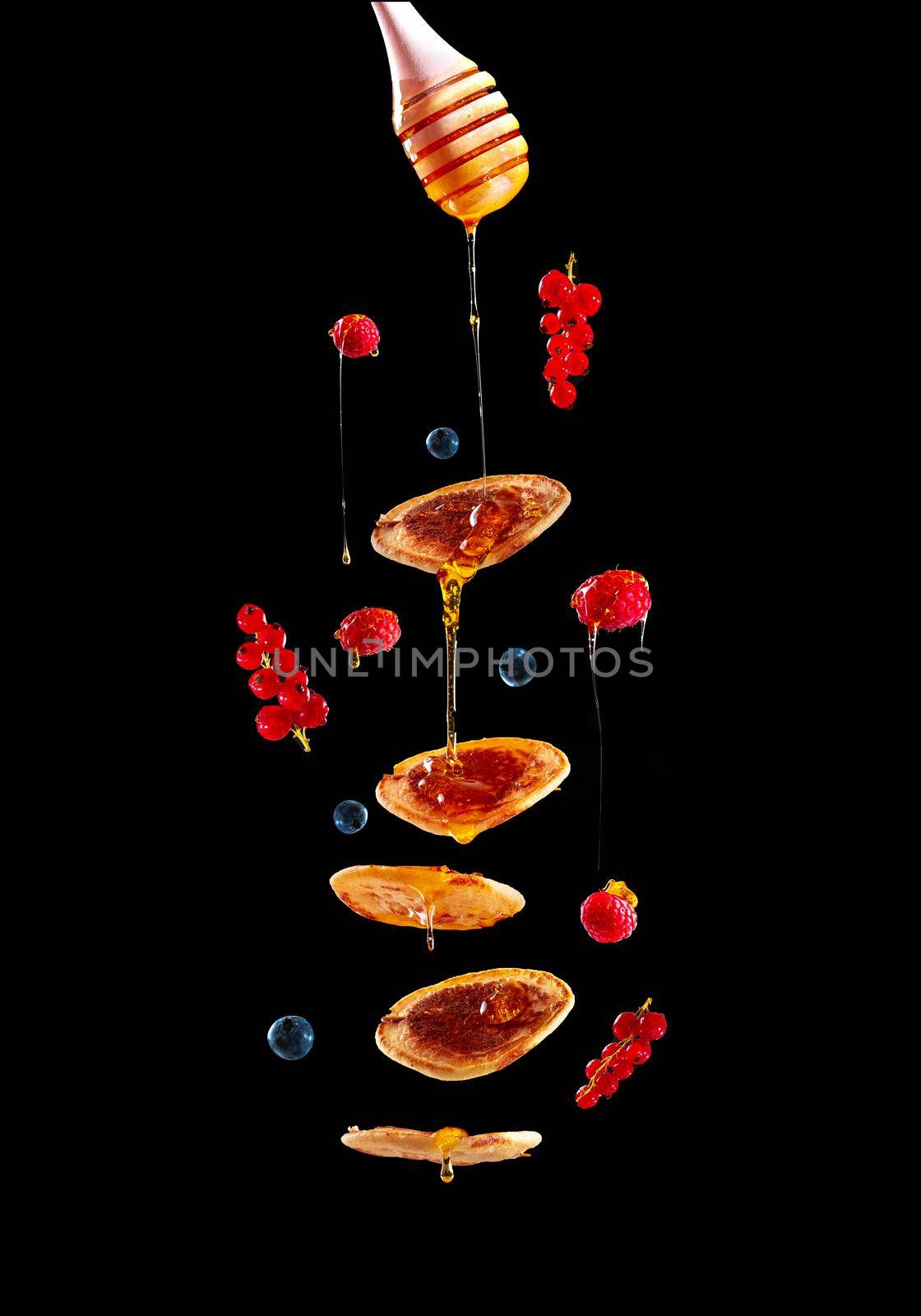 Pancakes with fruit and honey on a black background flying in the air. Mini pancakes with blueberries and raspberries. American breakfast on a dark background. Healthy eating for the whole family. Homemade pancakes with berries. Pancakes with honey. Homemade pancakes on a white background. European breakfast pancakes and berries