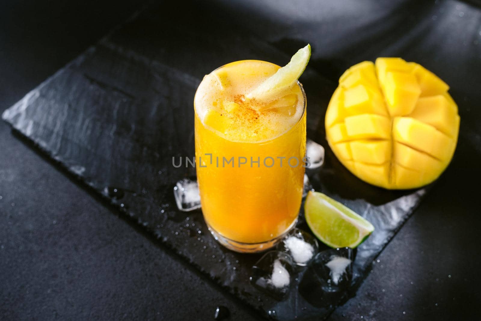 Mango juice on a dark background. Summer drink with ice and slices of mango and lime. Mango and lime juice nectar. A refreshing summer drink on a black background. Bright yellow and black colors