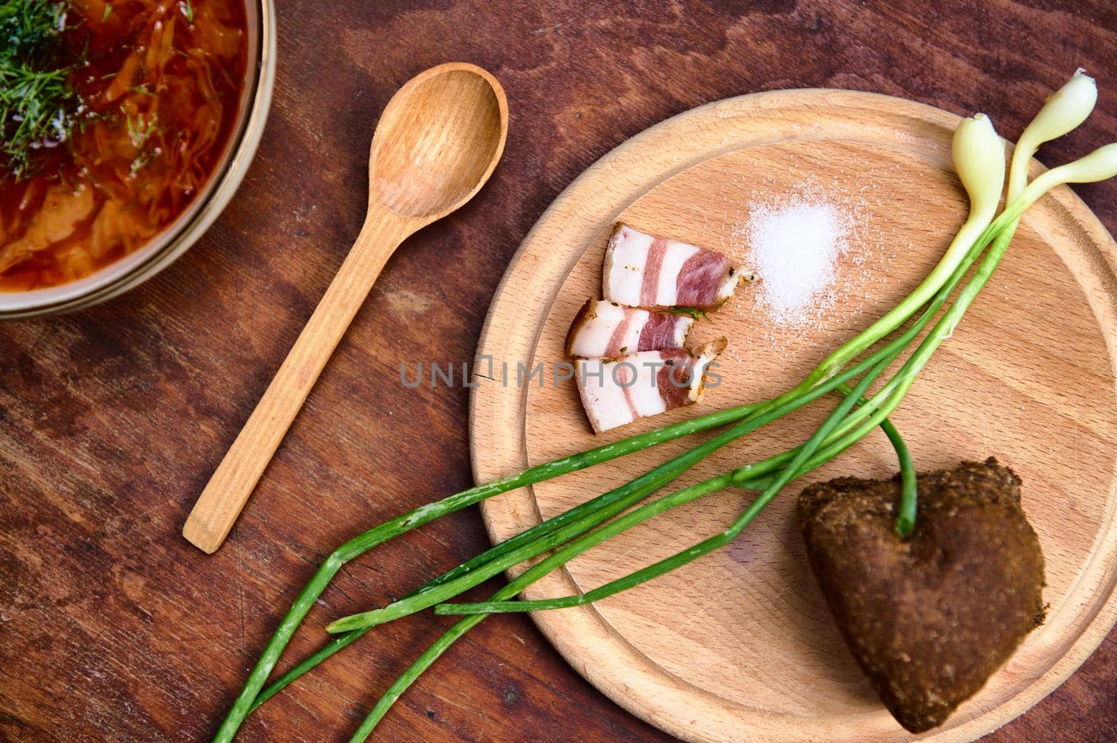 Top view of a wooden cutting board with a sliced bacon, salt, bread and green onion near a soup spoon and partial view of a red beetroot soup- traditional Ukrainian dish- Borscht on a wooden surface