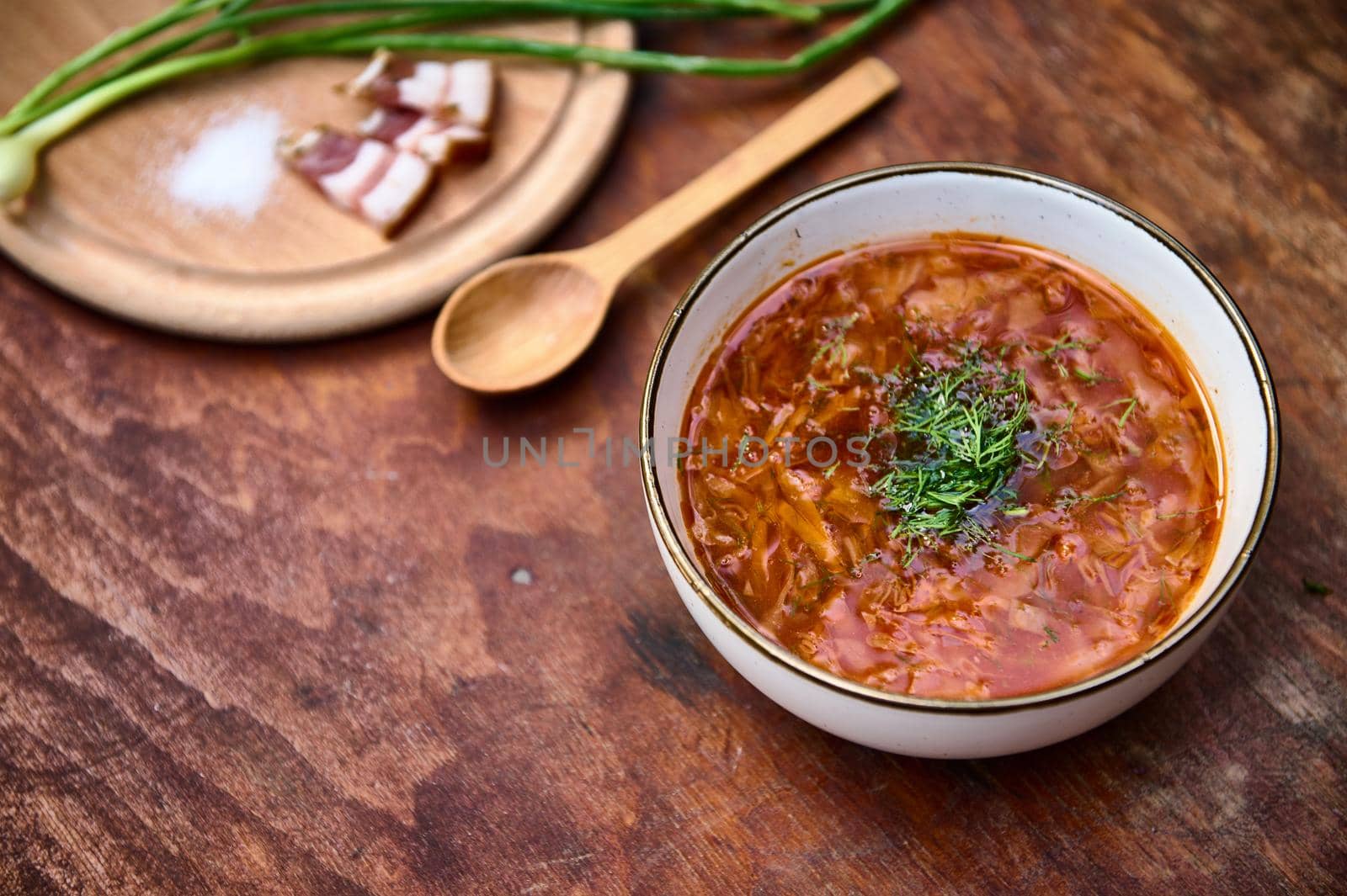 Ukrainian national dish: red beetroot soup - traditional tasty Borscht with parsley and dill, chopped bacon, salt and green onions nearby on a wooden board against a wood table background