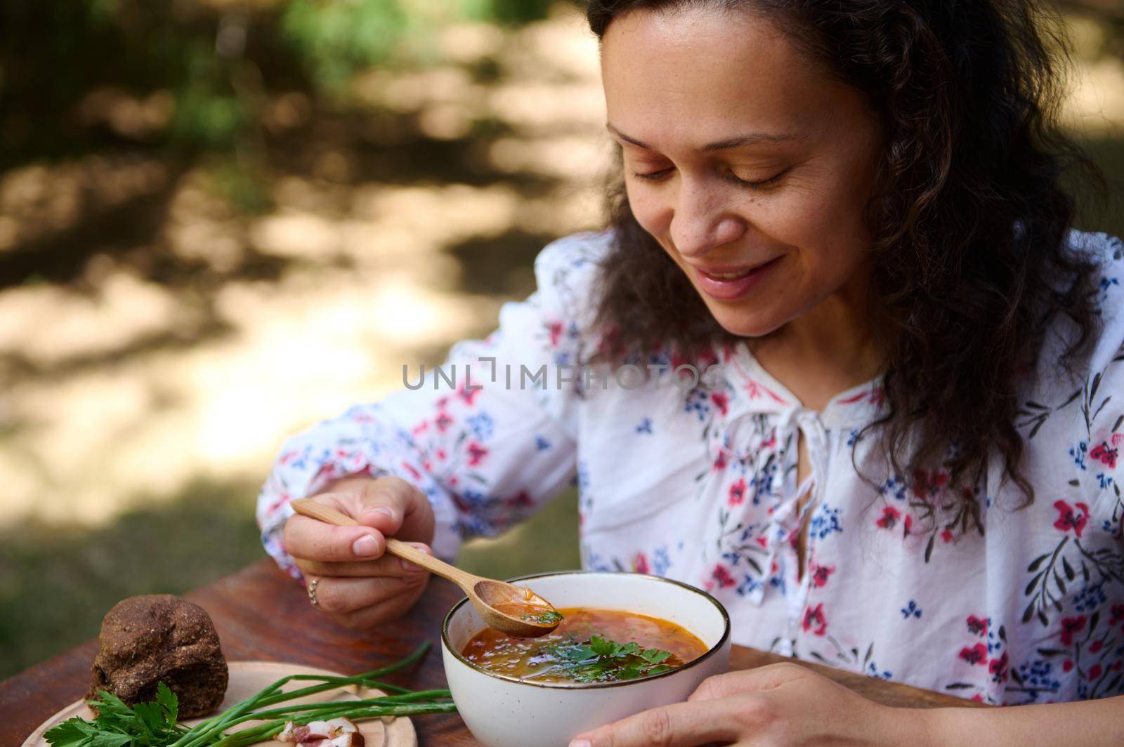 Close-up of a middle-aged charming multi-ethnic woman eating delicious traditional Ukrainian borscht - beetroot soup with whole grain rye bread and green onion outdoors. Slavic cooking
