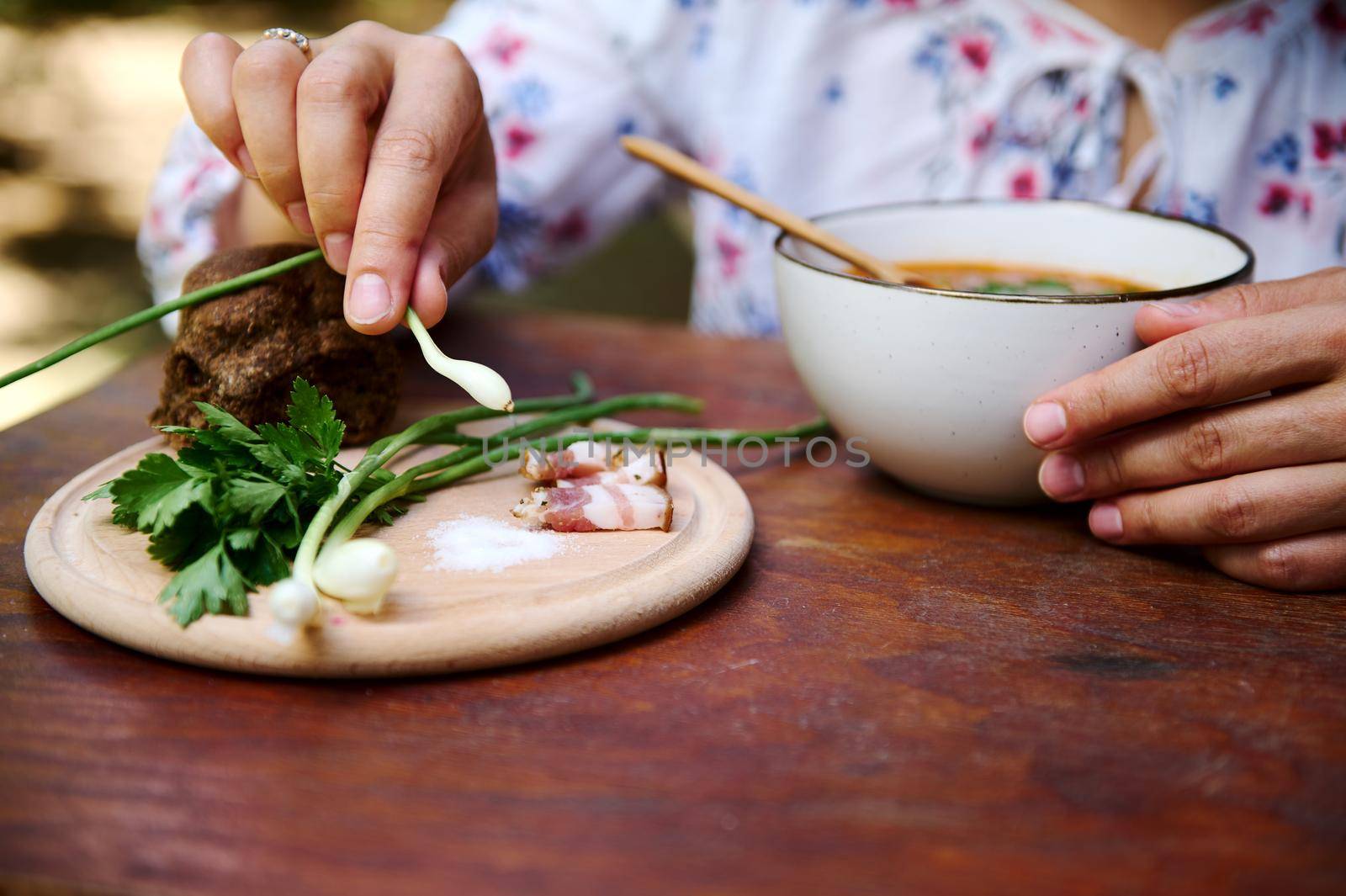 Close-up of a woman holding green onion while tasting Ukrainian national Borscht on a served rustic wooden table. Slavic traditional culinary. Red beetroot soup, sliced bacon, parsley on cutting board