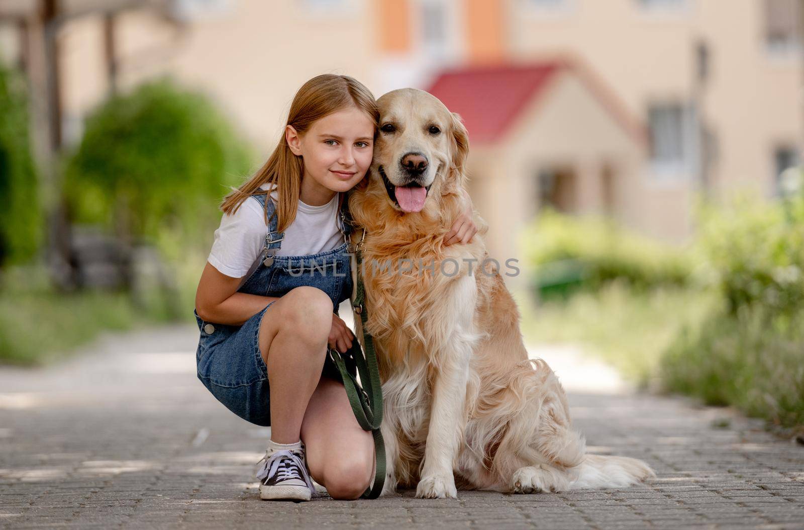 Preteen girl wearing hat hugging golden retriever dog sitting outdoors in summertime. Pretty kid petting fluffy doggy pet in city