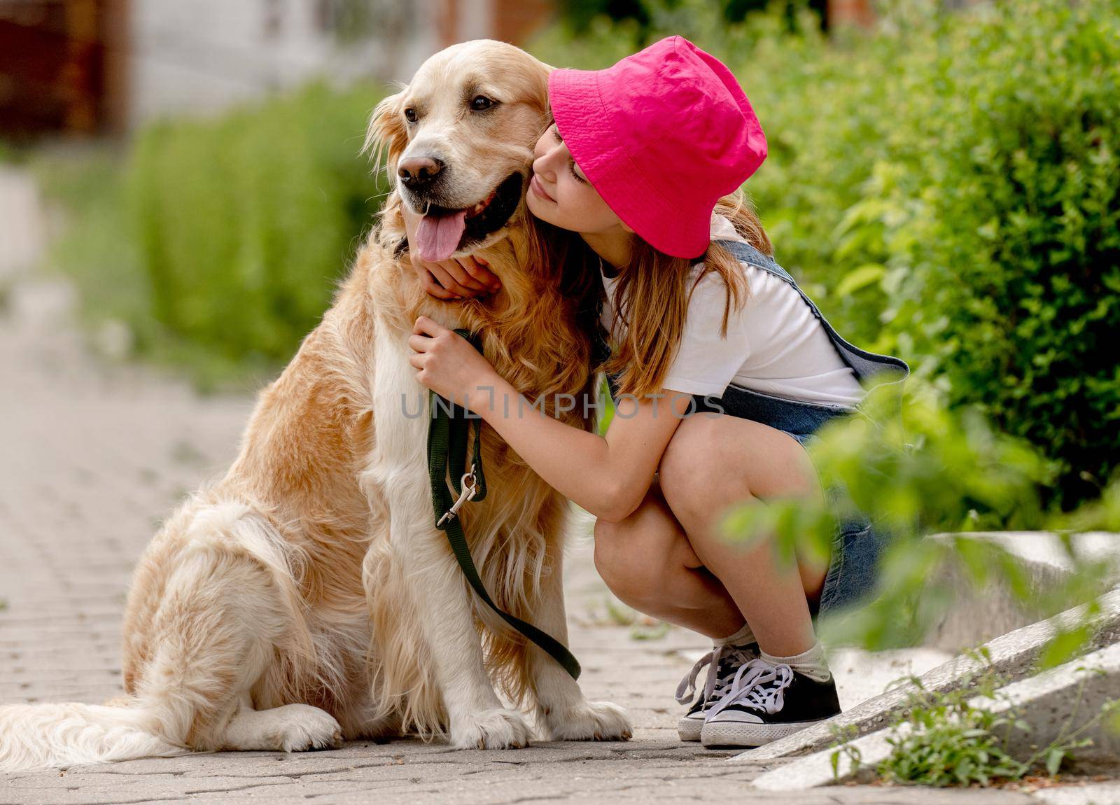 Preteen girl wearing hat hugging golden retriever dog sitting outdoors in summertime. Pretty kid petting fluffy doggy pet in city