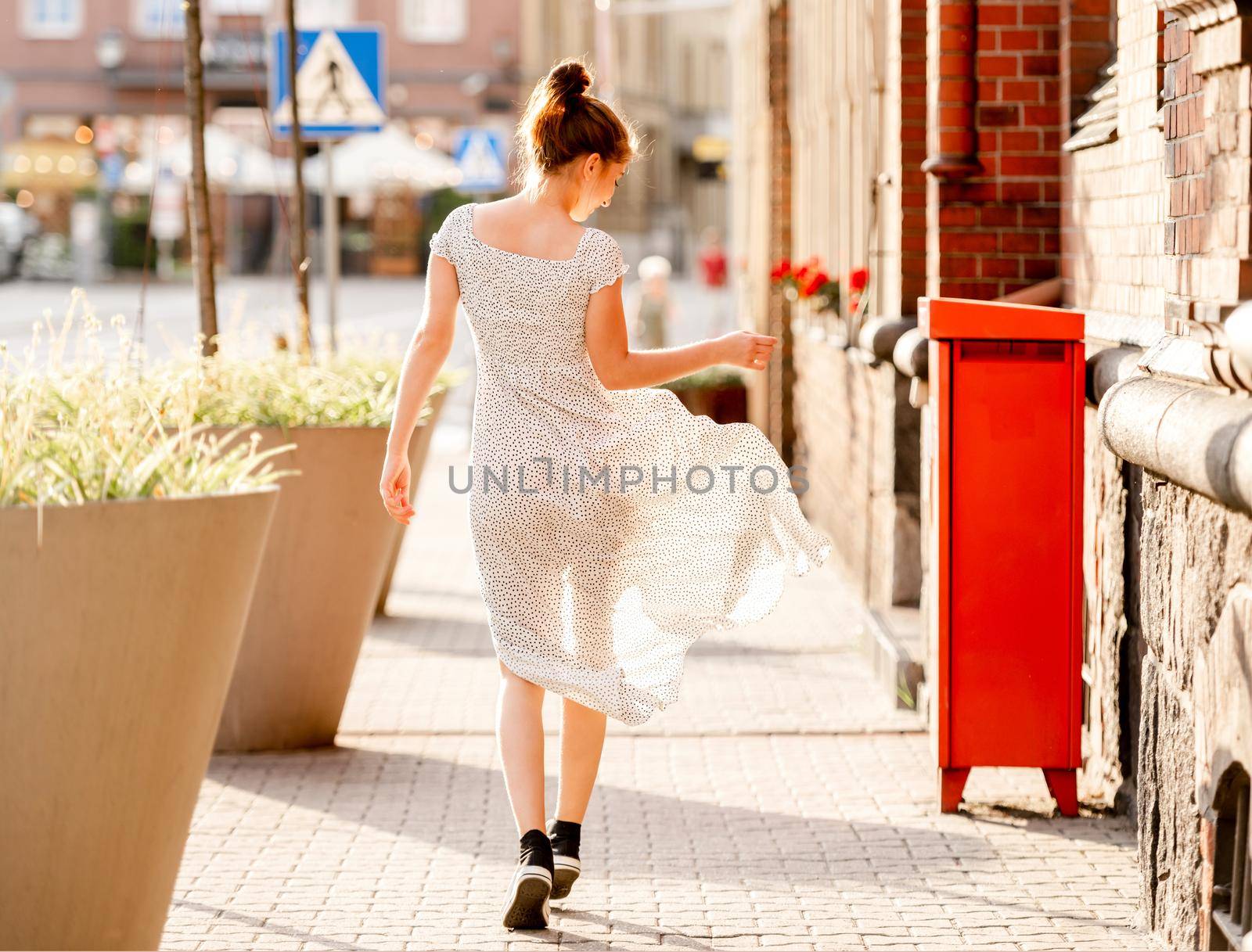 Beautiful girl in long dress walking at street in city with ancient architecture, view from back. Pretty female teenager outdoors in summer time