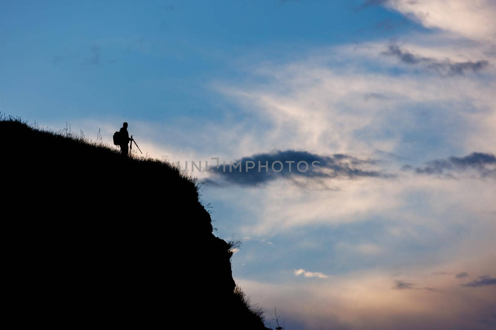 silhouette of unrecognizable photographer with tripod on cliff with evening blue sky with darkening clouds background