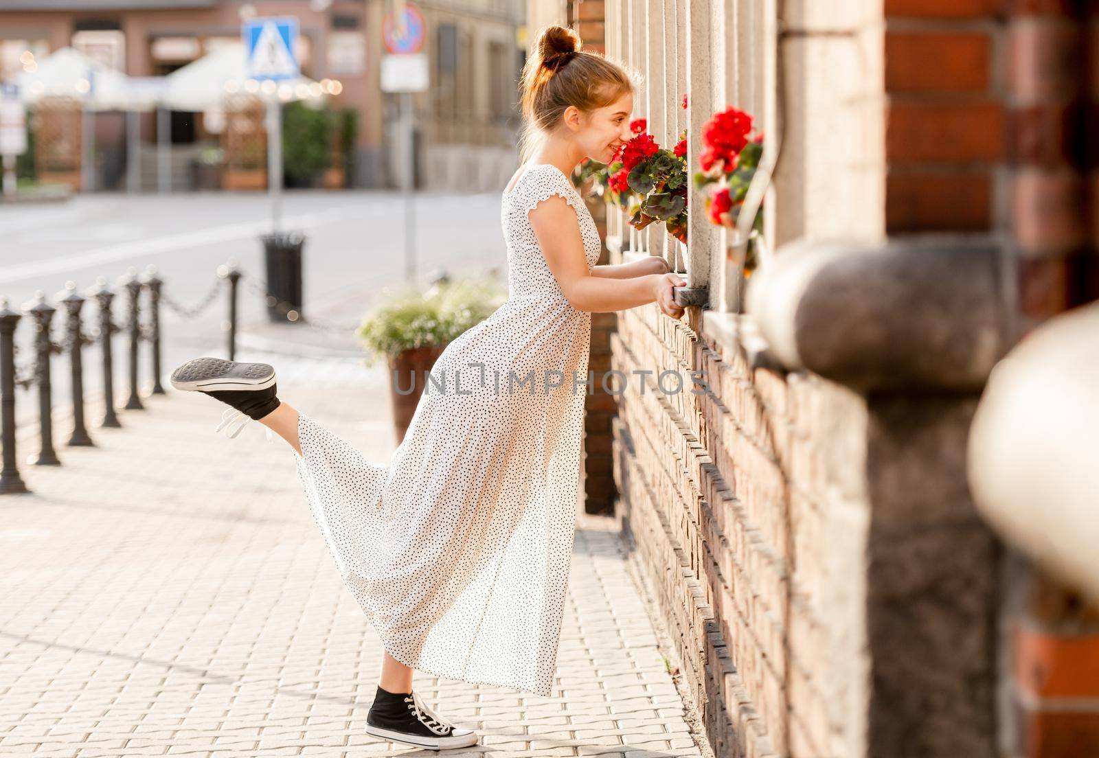 Beautiful girl in long dress sniffing red flowers at street in city with ancient architecture. Pretty female teenager model outdoors in summer time
