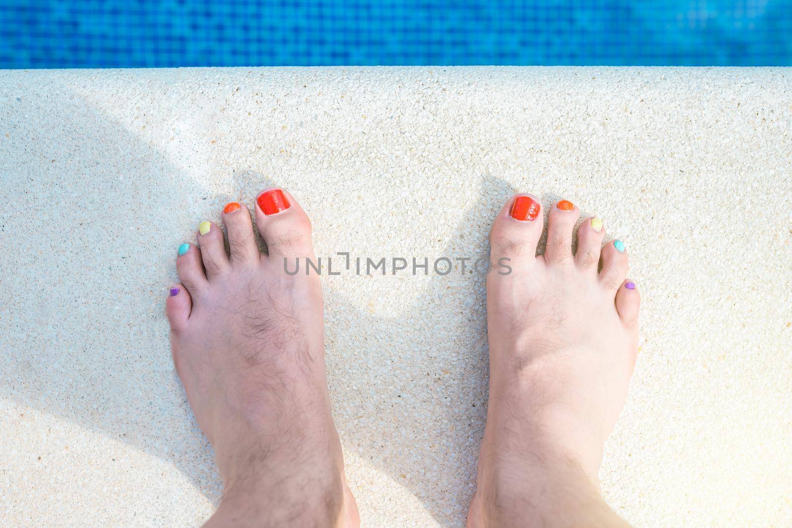 feet of a hairy man with his nails painted in LGBTIQ colours. bare feet at the edge of a swimming pool. concept of holidays and diversity. outdoors, natural light, summer days.