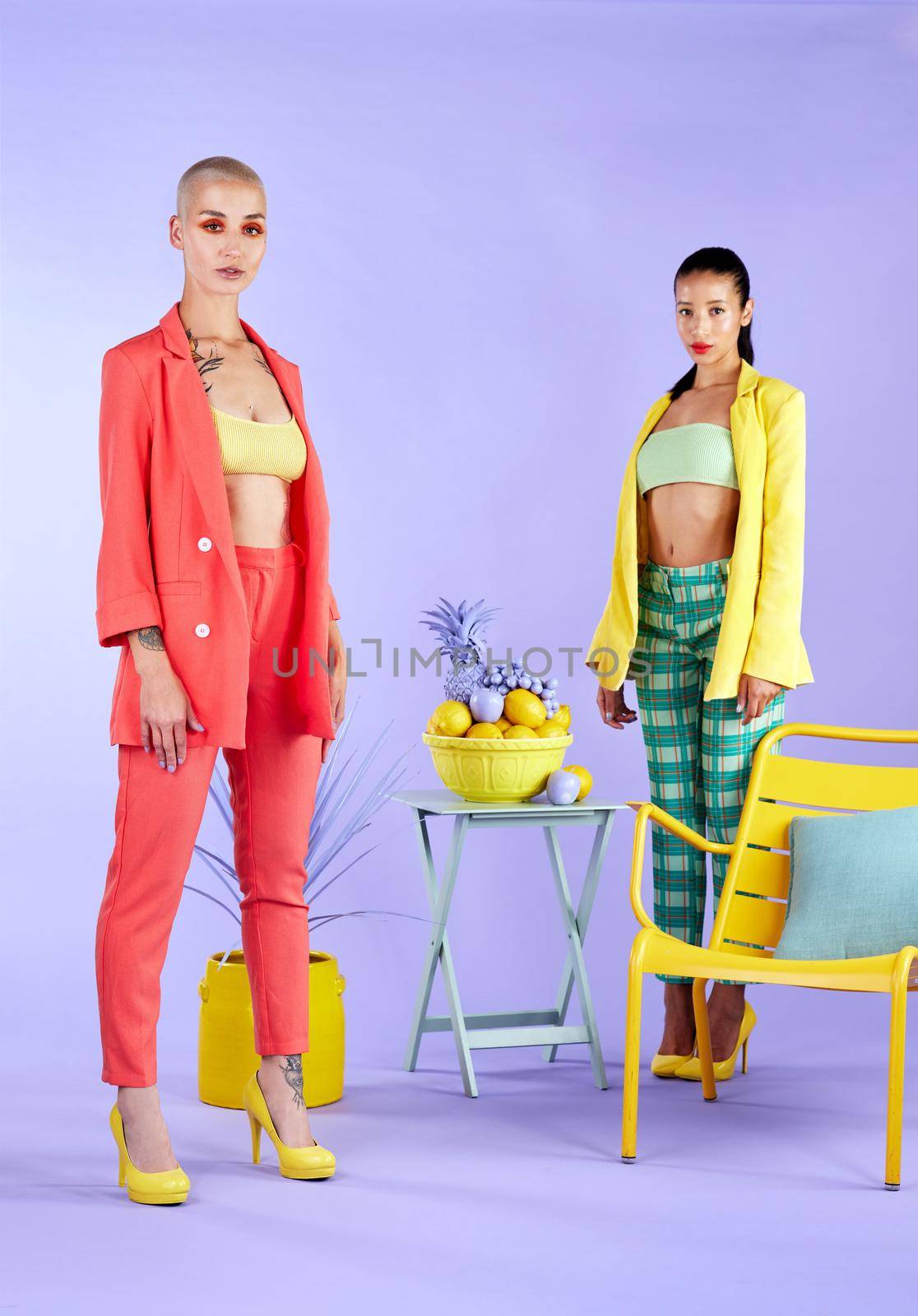 Full length studio portrait of two attractive young women dressed in funky attire against a purple background.