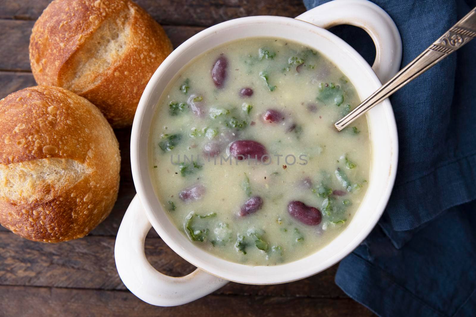 Portuguese Caldo Verde soup flat lay with bread, napikin and spoon in bowl, on dark wooden table.