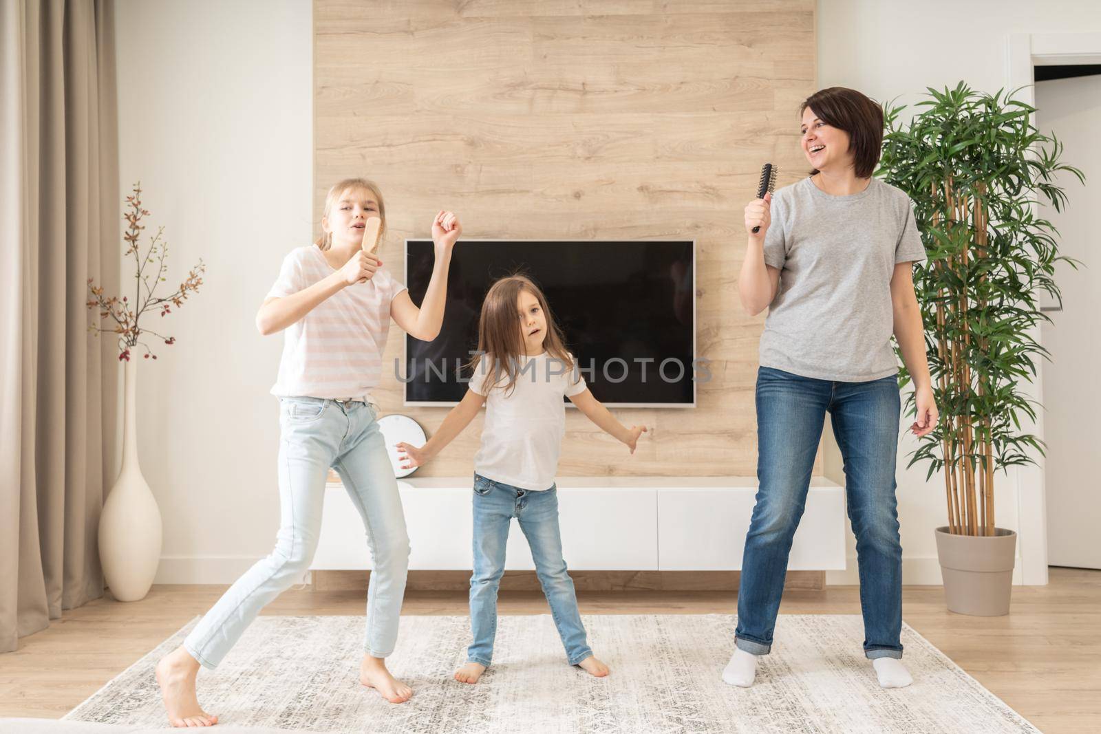 Happy mother and two daughters having fun singing karaoke song in hairbrushes. mother laughing enjoying funny lifestyle activity with teenage girl at home together. by Mariakray