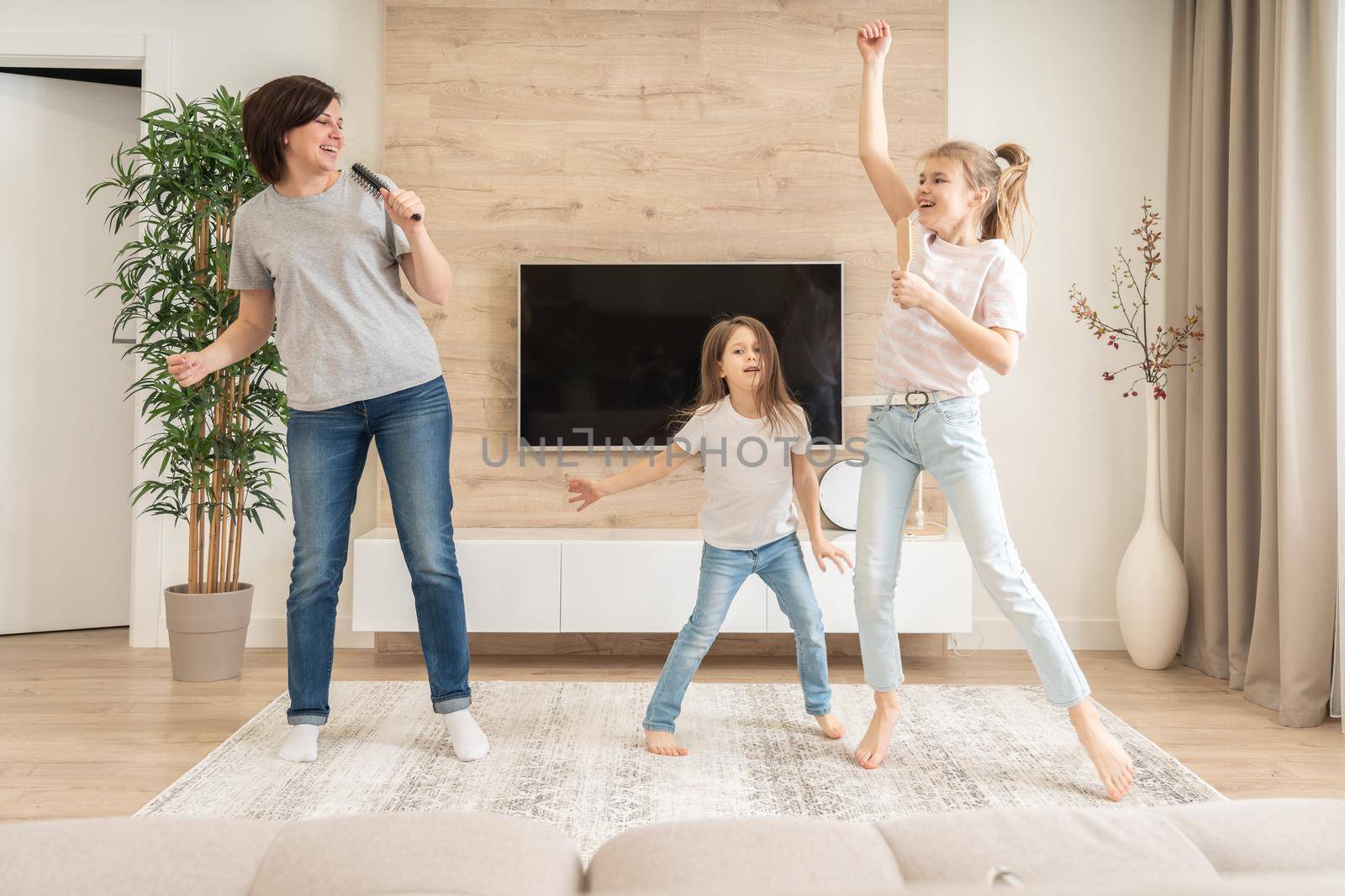 Happy mother and two daughters having fun singing karaoke song in hairbrushes. mother laughing enjoying funny lifestyle activity with teenage girl at home together. by Mariakray