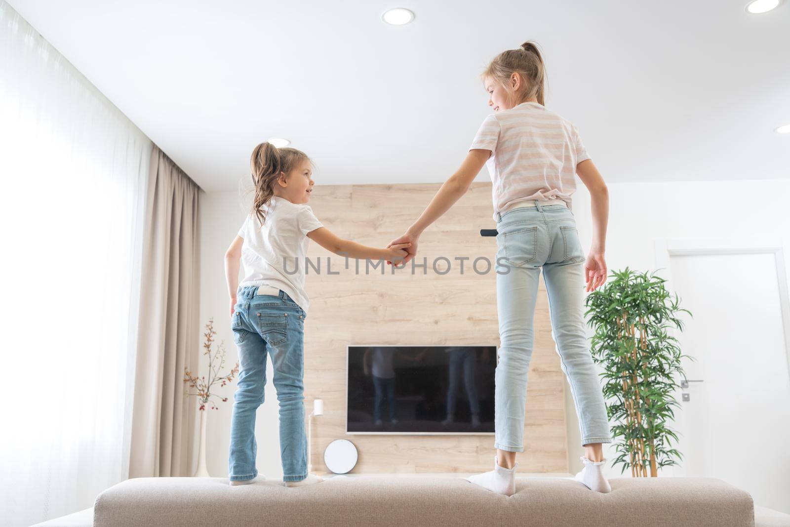 Two happy sisters jumping on couch in living room having fun