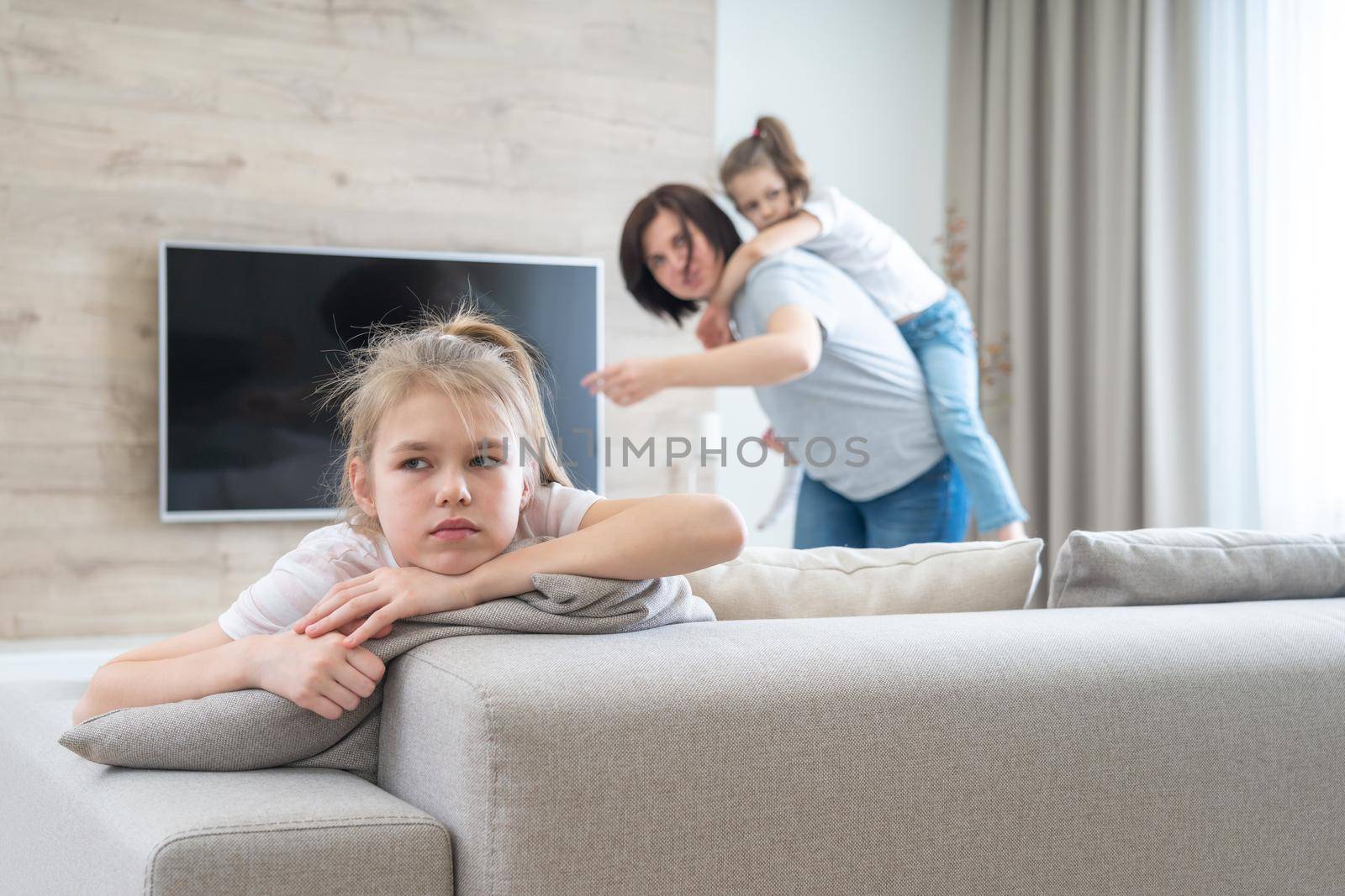 Preteenage sad girl sitting on a couch while mother having fun with sister, jealousy concept by Mariakray