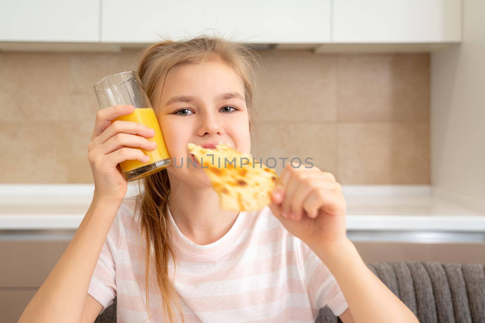Teenage girl eating a slice of pizza and drinking orange juice in the kitchen by Mariakray
