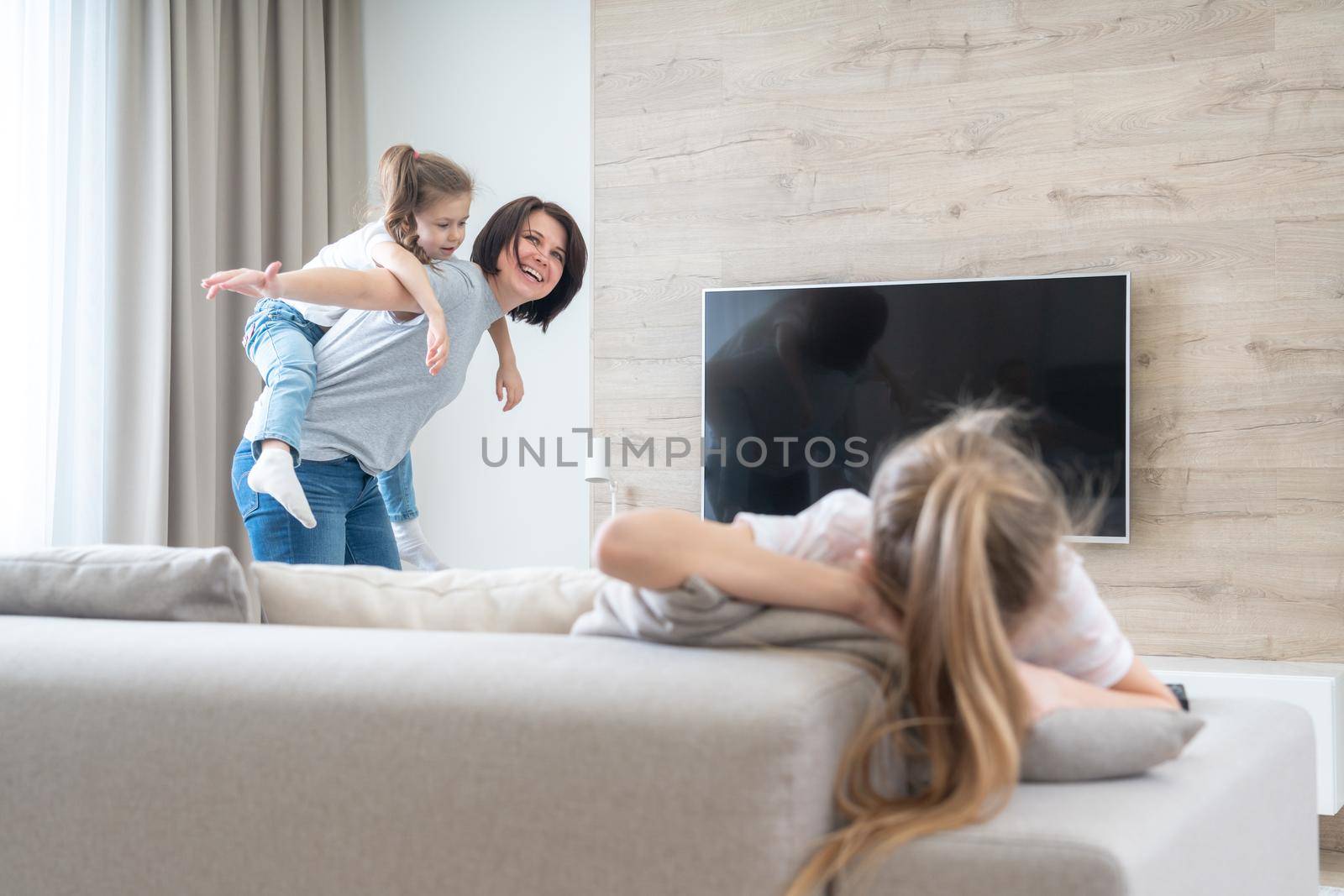 Preteenage sad girl sitting on a couch while mother having fun with sister, jealousy concept by Mariakray
