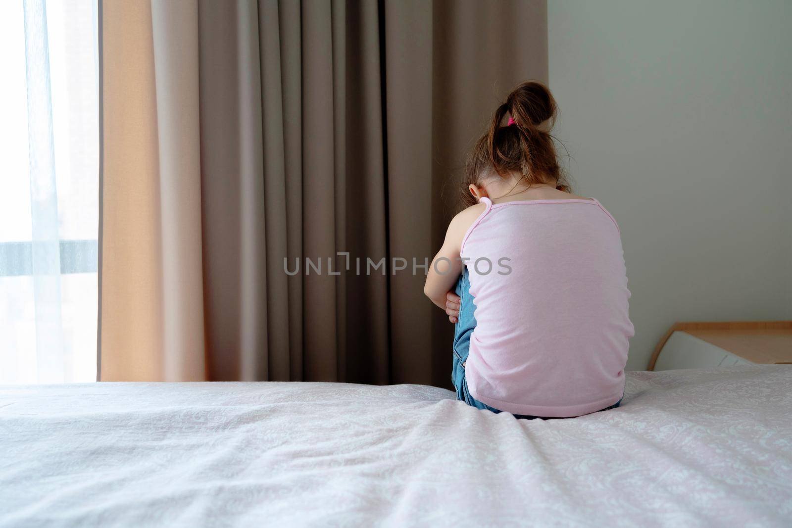 A little sad girl sitting on a bed in bedroom, back view by Mariakray